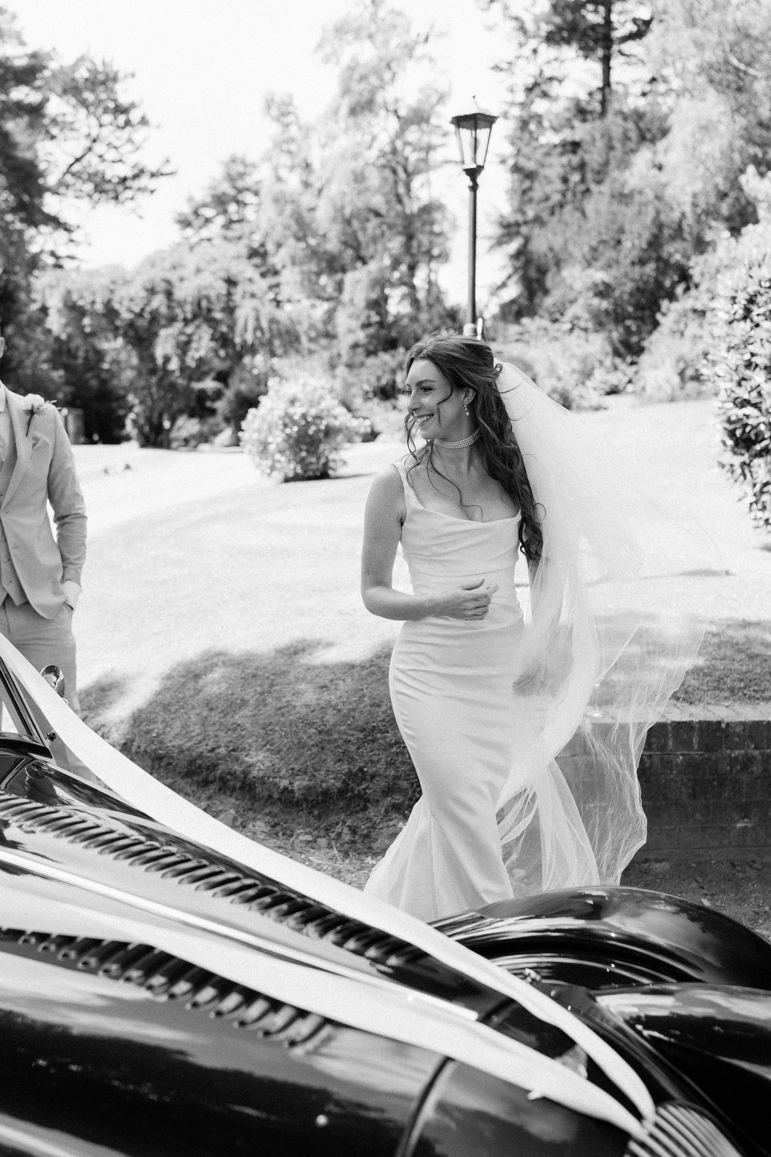 Smiling bride in fitted white gown and flowing veil walking beside a vintage car on a sunny wedding day.