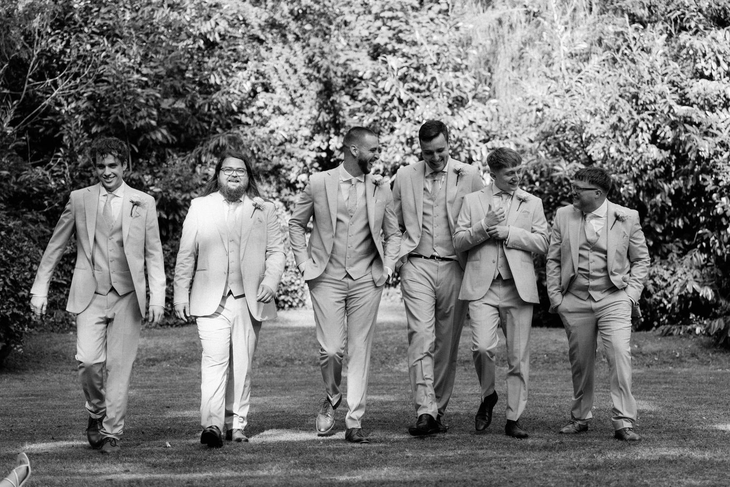 Groom and groomsmen walk together in matching light suits through a garden before the wedding.