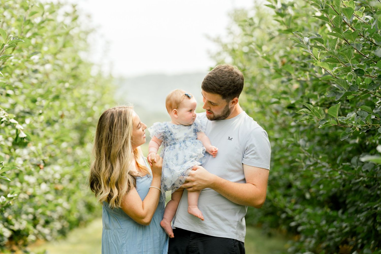 Young family with baby in lush green orchard, parents smiling at infant in fathers arms on a misty day.