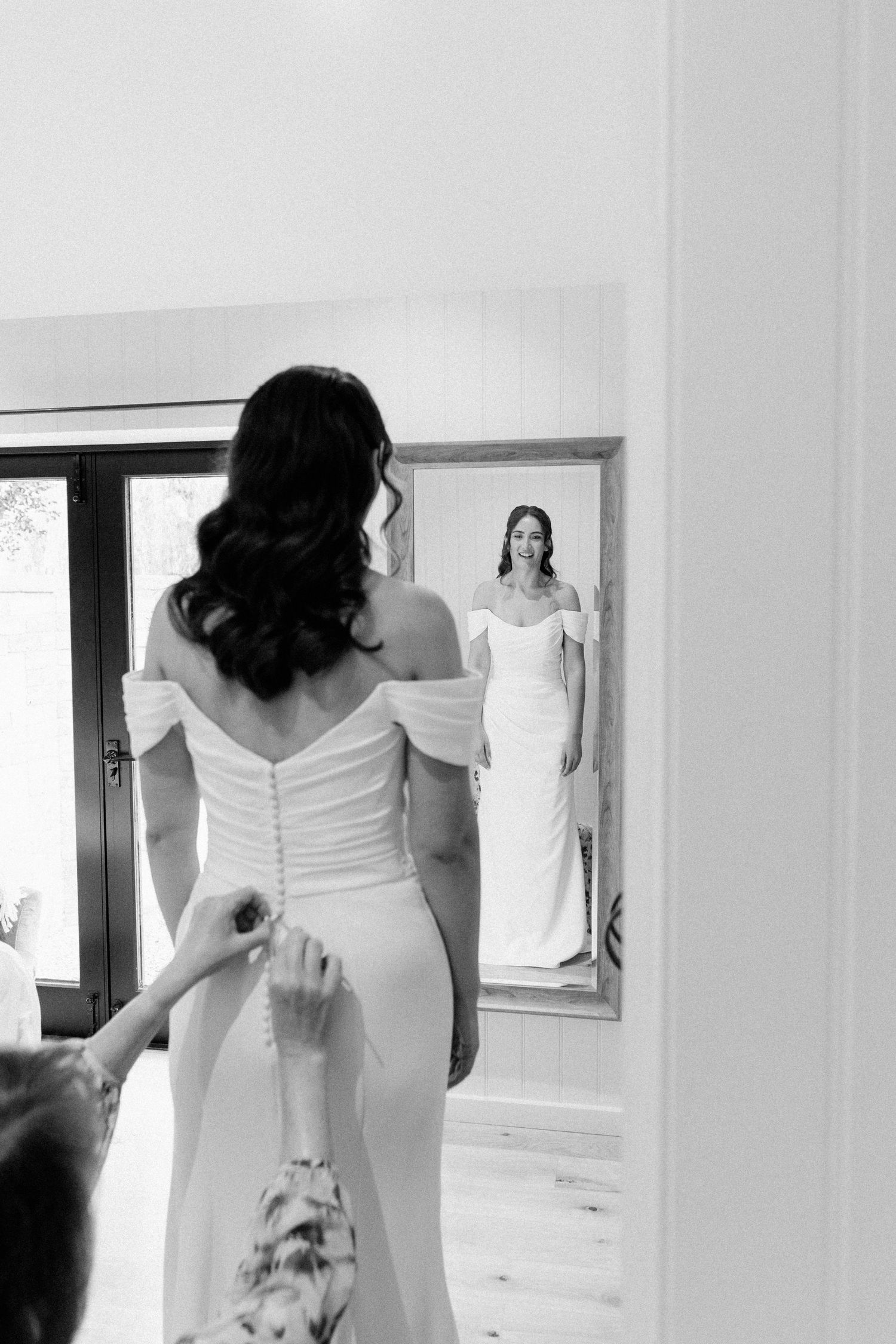 Bride in off-shoulder white gown views her reflection in mirror while bridesmaid fastens buttons, black and white photo.