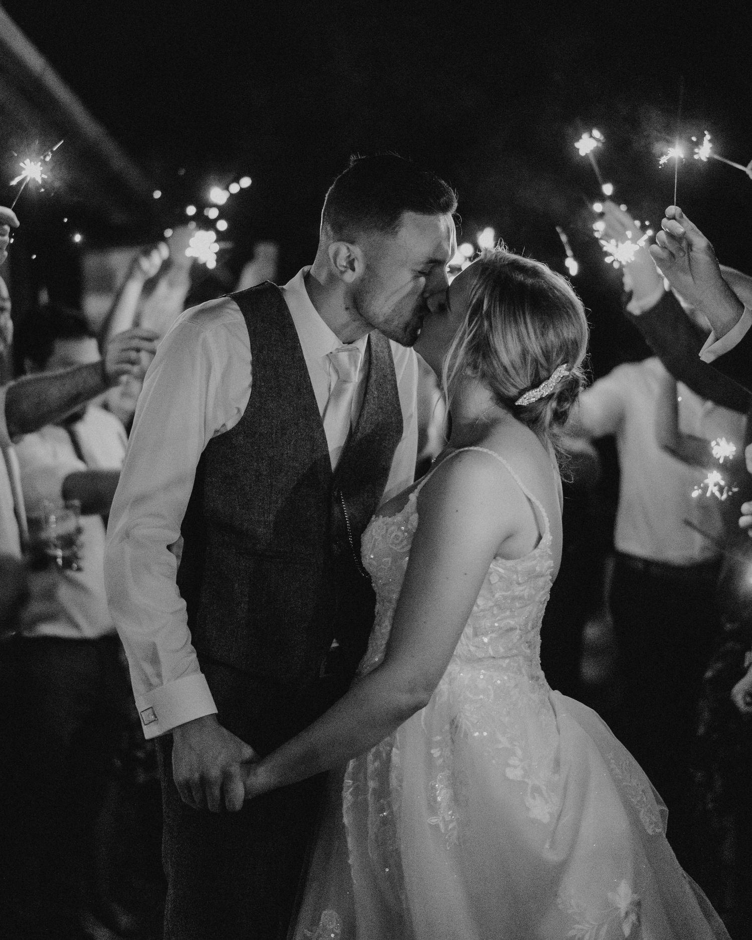 Black and white photo of a newlywed couple kissing during a sparkler send-off at their wedding reception.