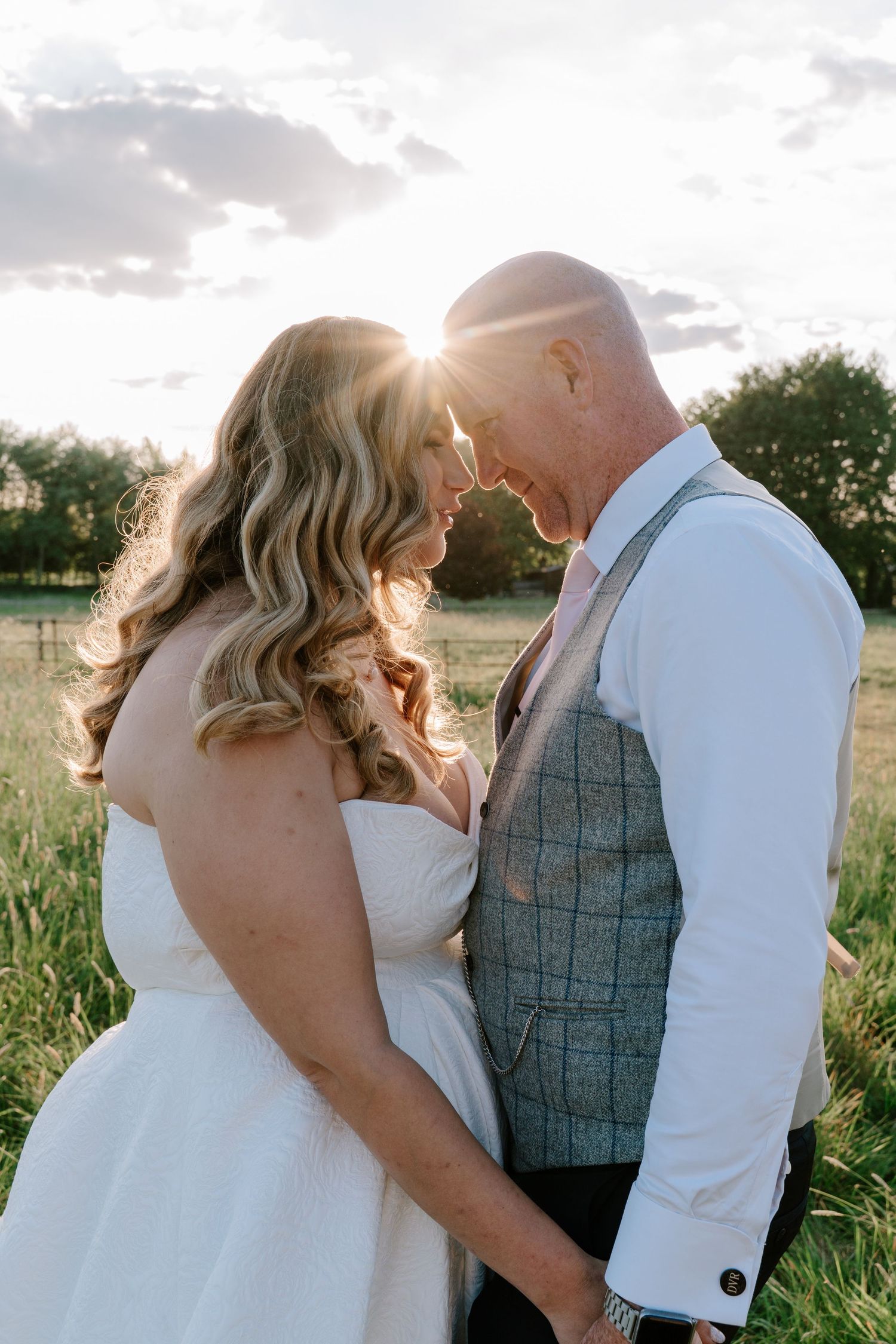 Bride and groom touch foreheads at golden sunset in a green field, sun flare glowing between them in romantic wedding portrait.