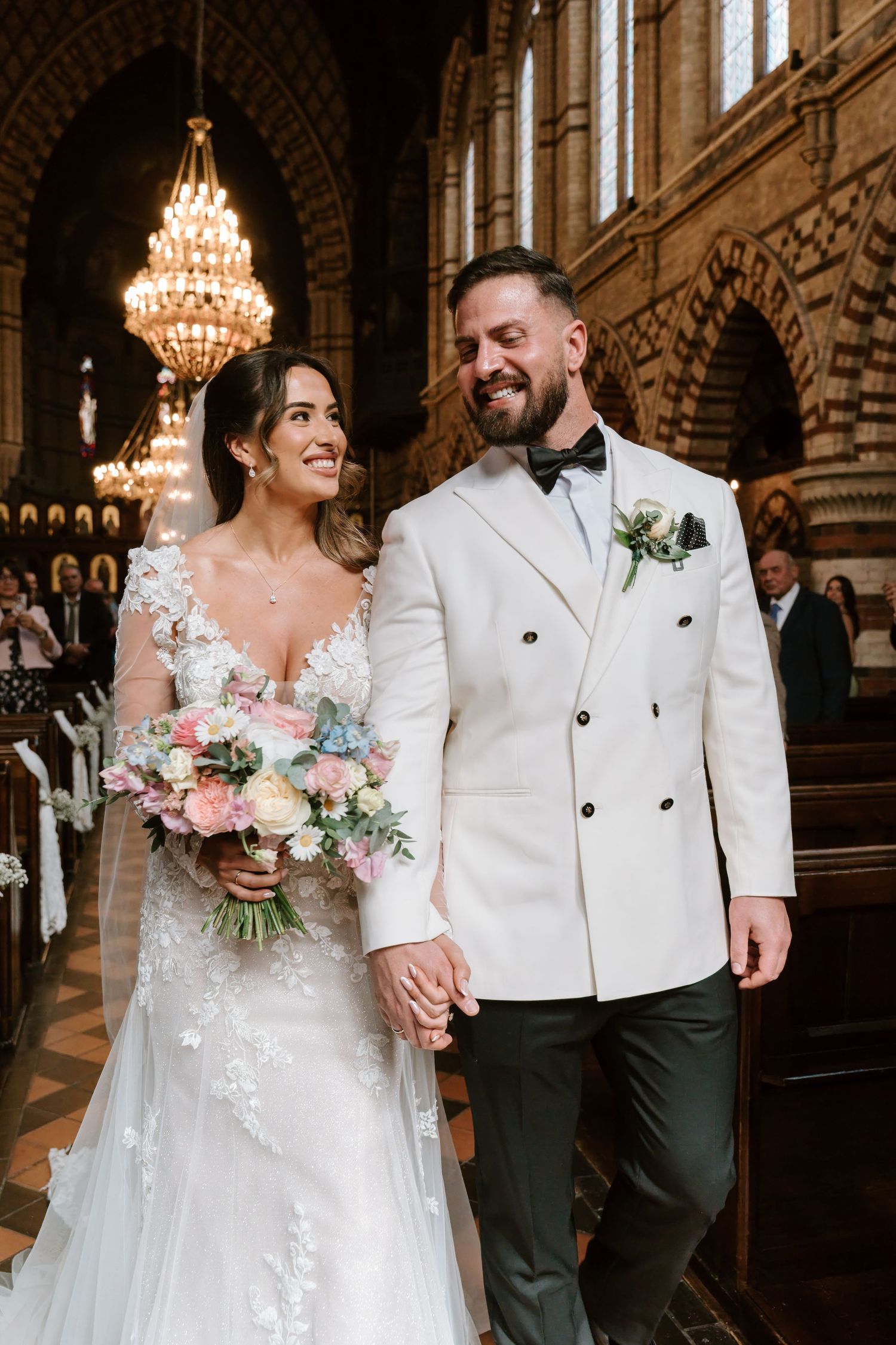 Bride and groom walk hand in hand down church aisle, she holds pastel bouquet, he wears white double-breasted jacket.