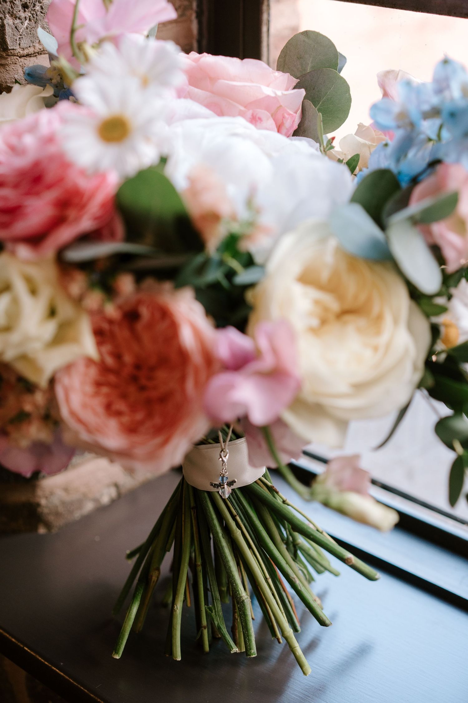 Elegant bridal bouquet with pink, white, and blue flowers including roses and daisies, tied with ribbon on a dark surface.