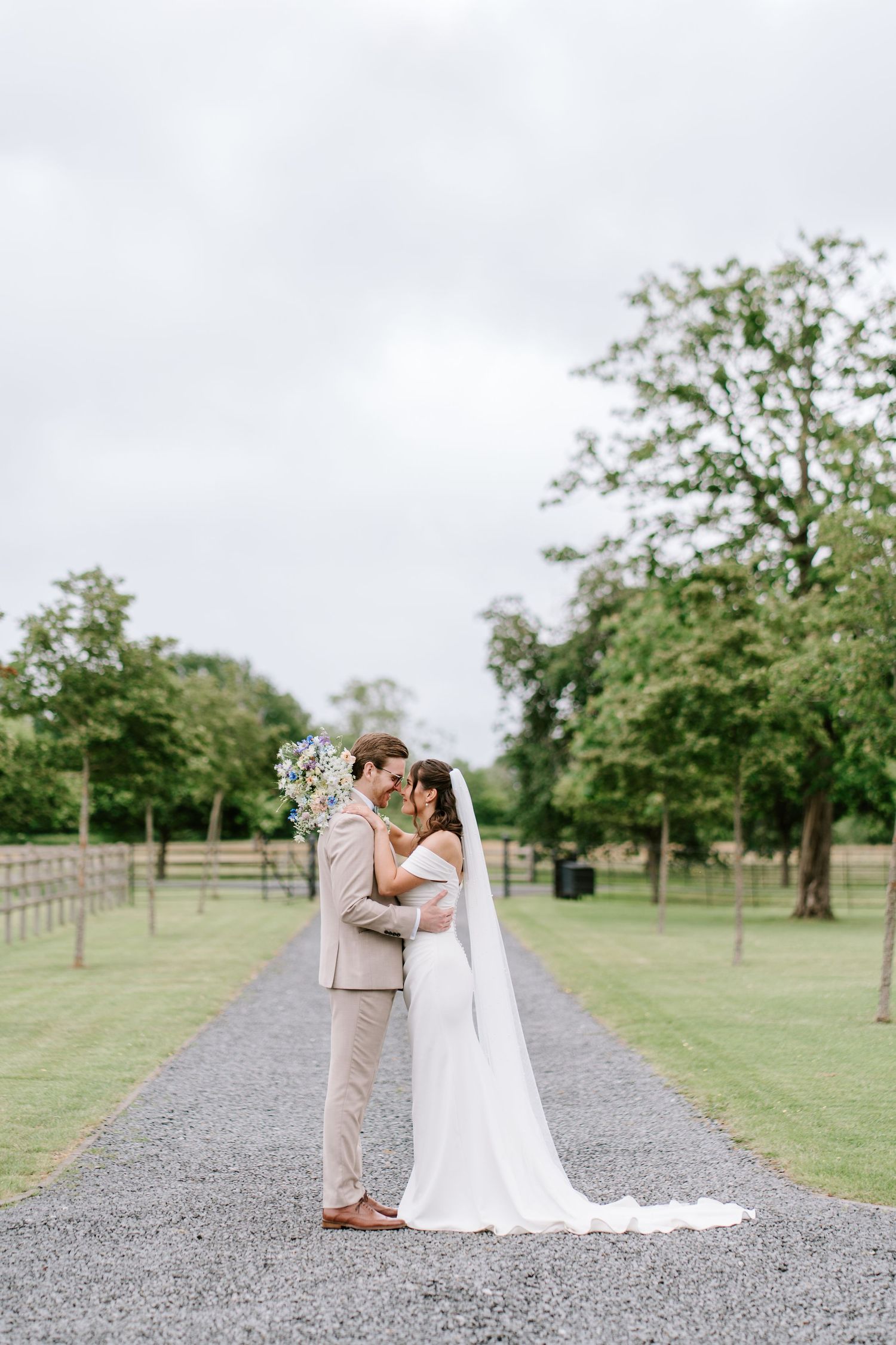 Bride and groom embrace on a gravel path surrounded by lush green trees at an elegant outdoor wedding venue.