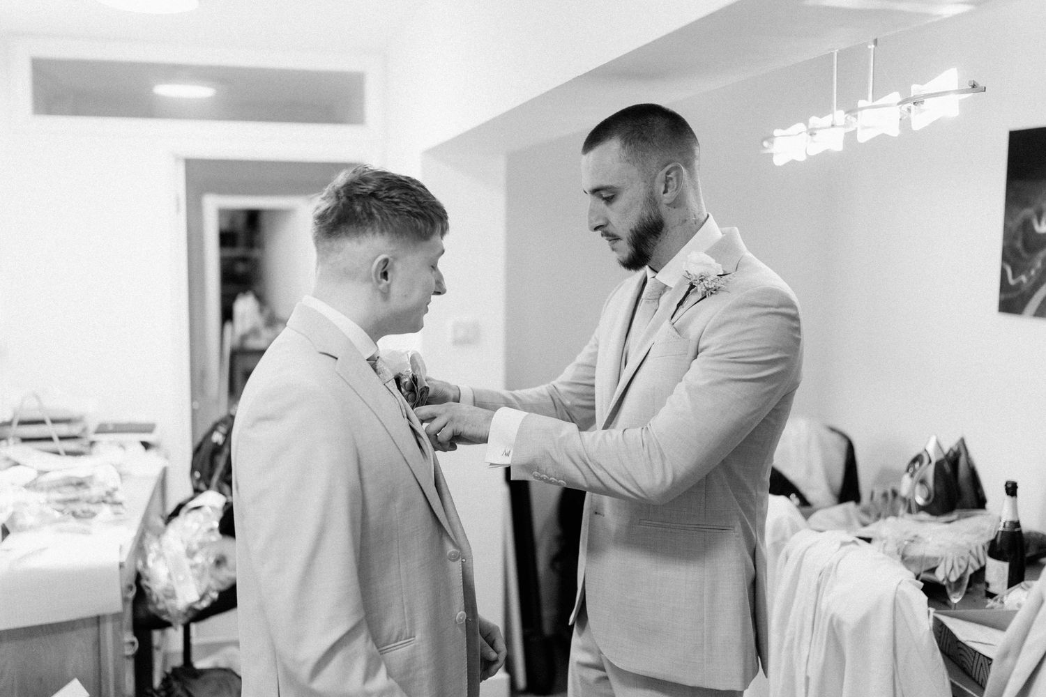 Groom adjusting boutonniere on groomsman in suits during wedding preparations in black and white.
