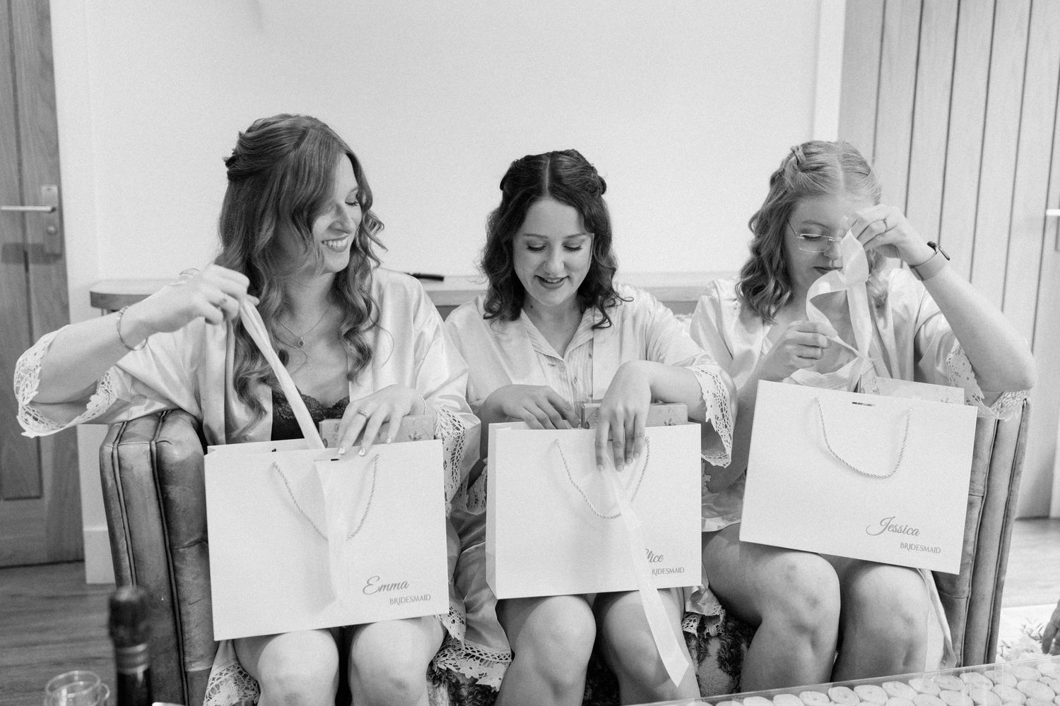 Three bridesmaids in robes joyfully opening personalized gift bags while seated together before a wedding ceremony.
