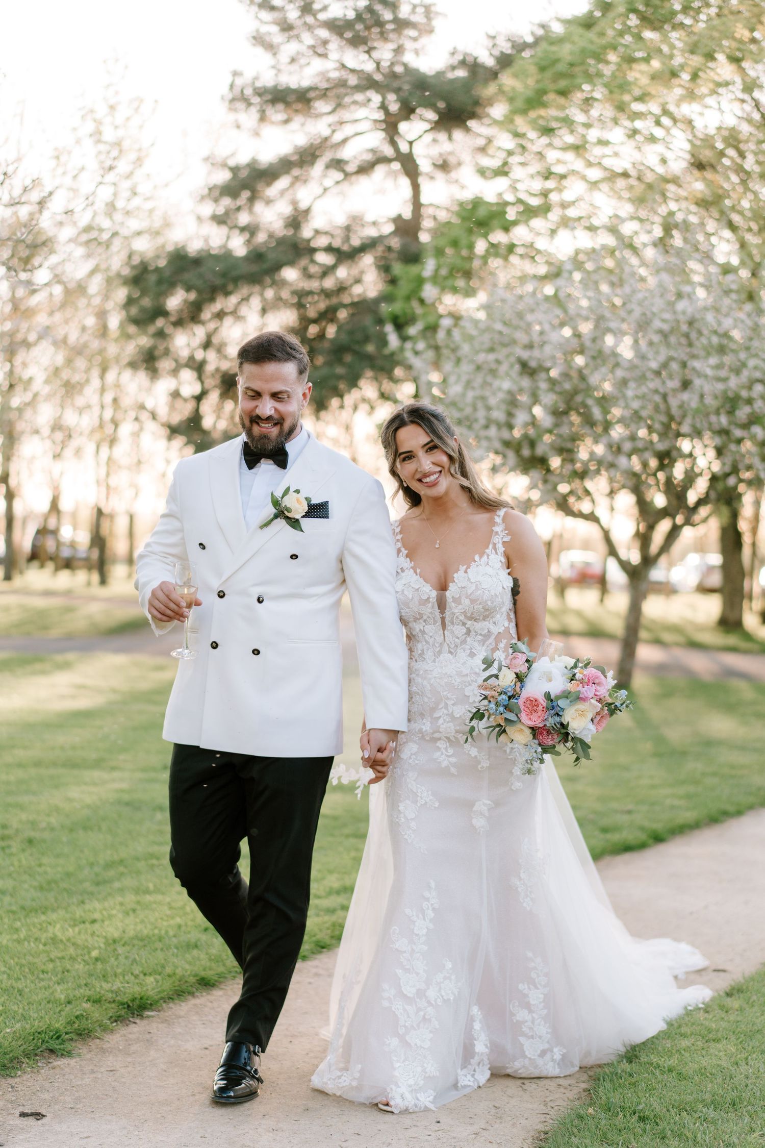 Bride and groom walk hand-in-hand outdoors, she in lace gown with bouquet, he in white tuxedo jacket.