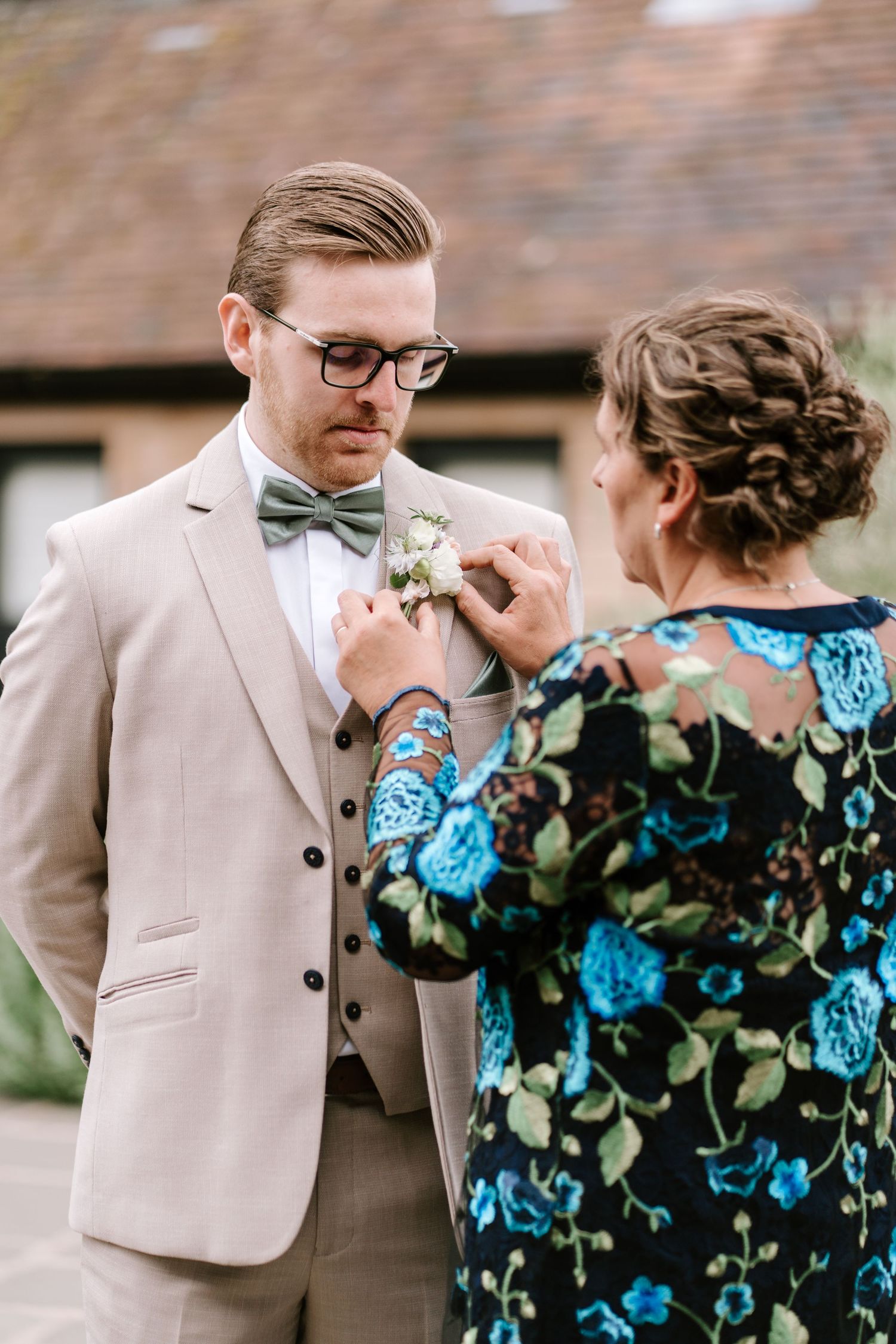 Groom in beige suit and bow tie having boutonniere pinned by woman in floral dress outdoors.