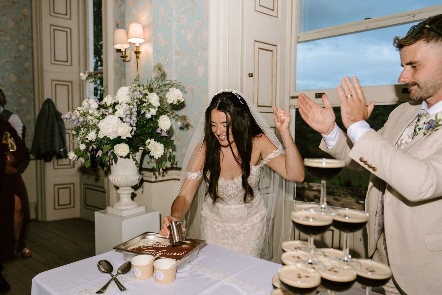 Bride and groom cutting wedding cake at elegant reception with white floral arrangements and dessert display.