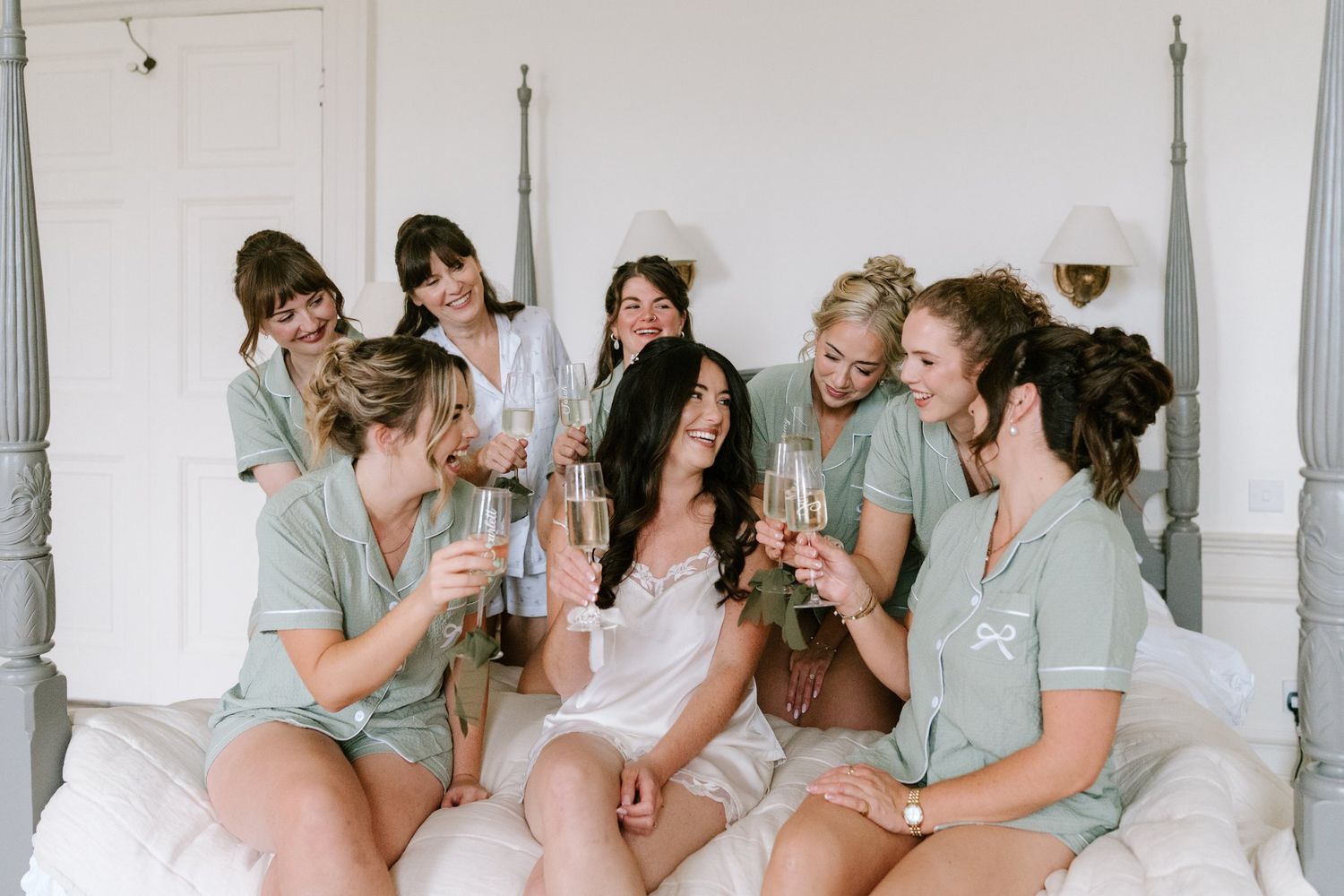 Bridesmaids in sage green pajamas toast champagne with bride in white lingerie on bed during wedding morning prep.