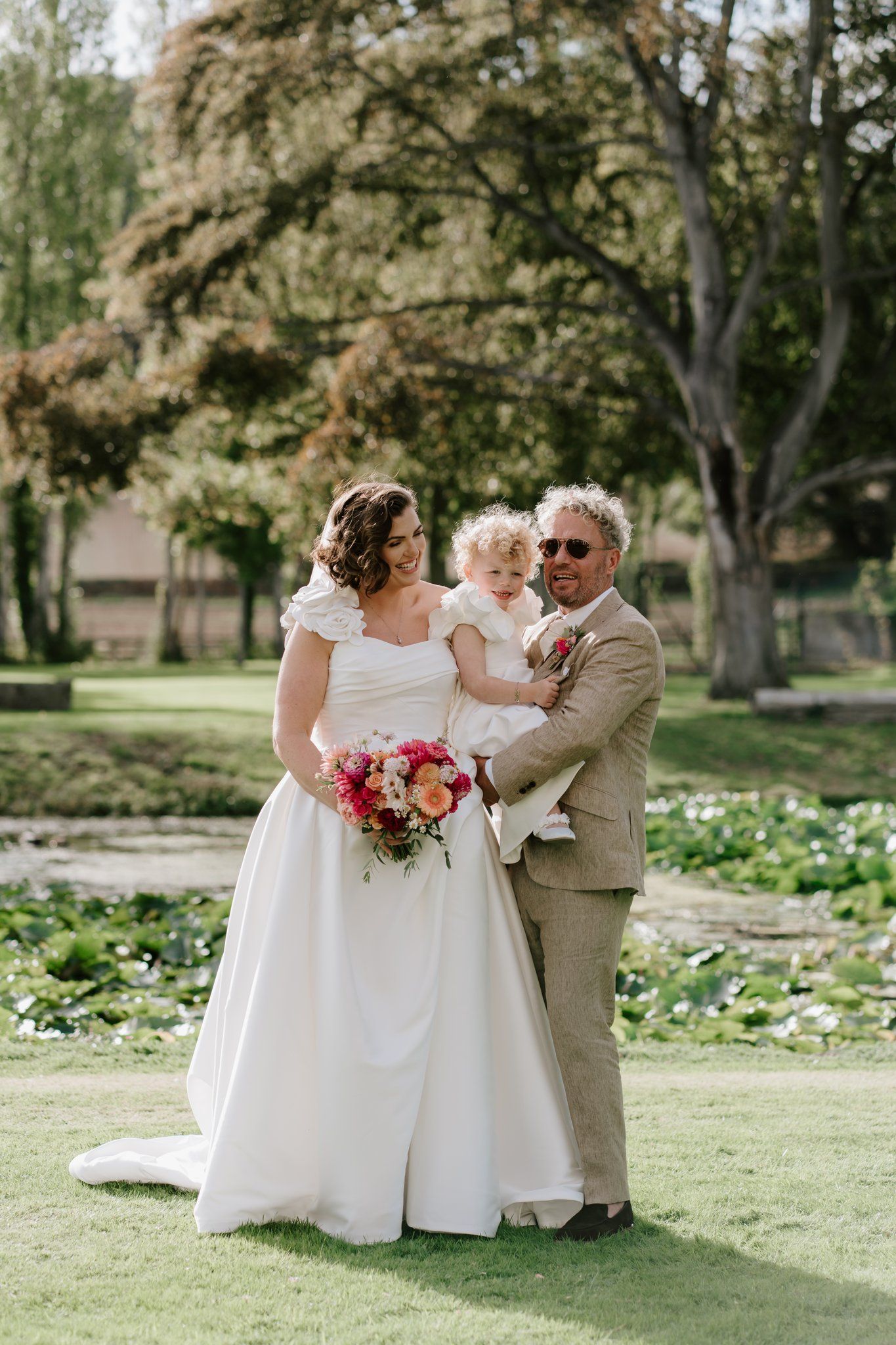 Bride in white gown and groom in tan suit holding toddler, posing outdoors in lush garden with colorful bridal bouquet.
