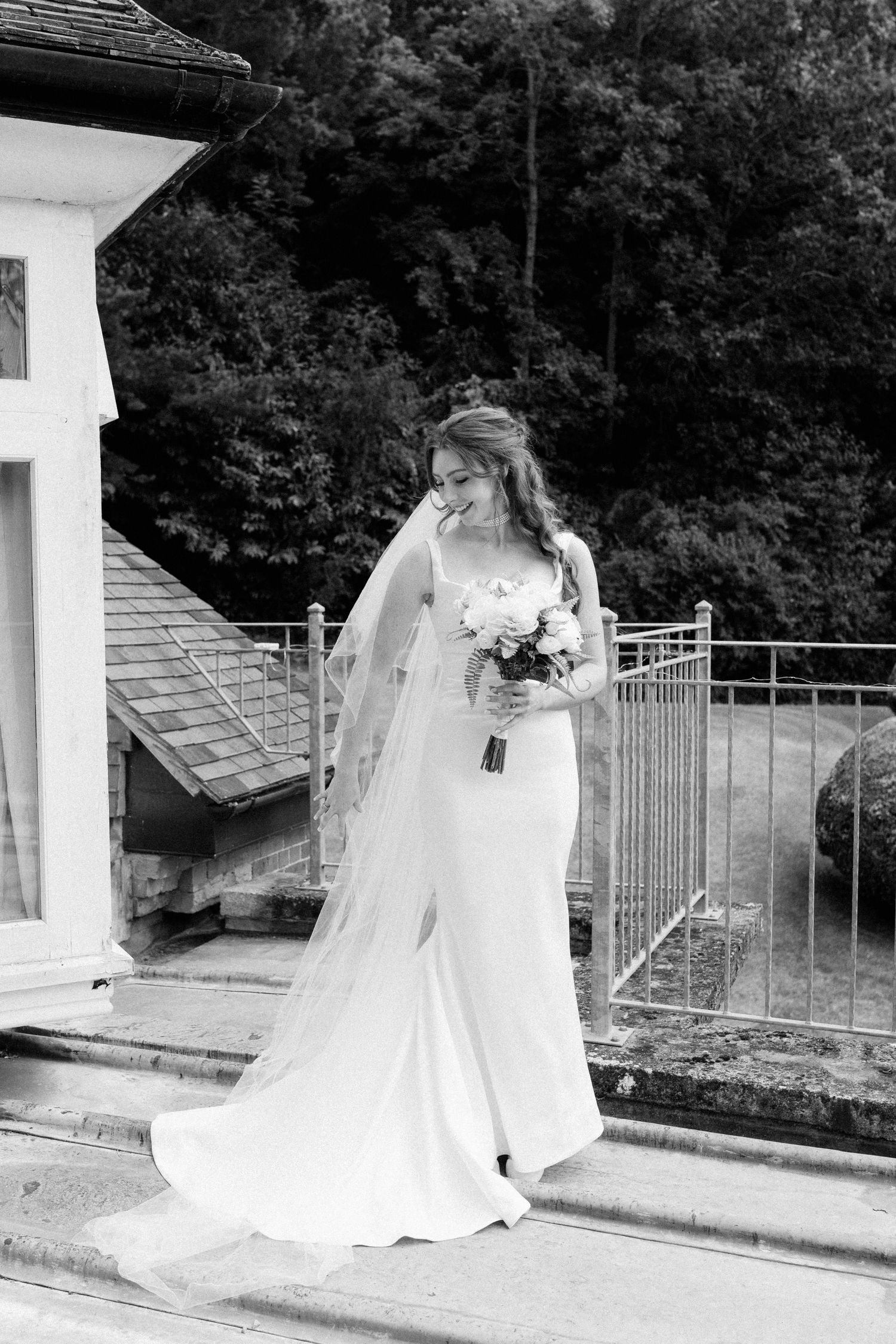 Bride in fitted white gown and long veil holding bouquet on outdoor steps in black and white wedding photo.