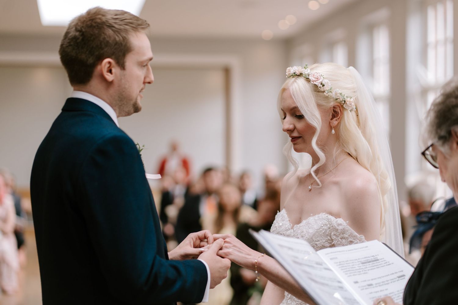 Groom placing wedding ring on bride's finger during ceremony, bride smiling in lace gown with floral headpiece.