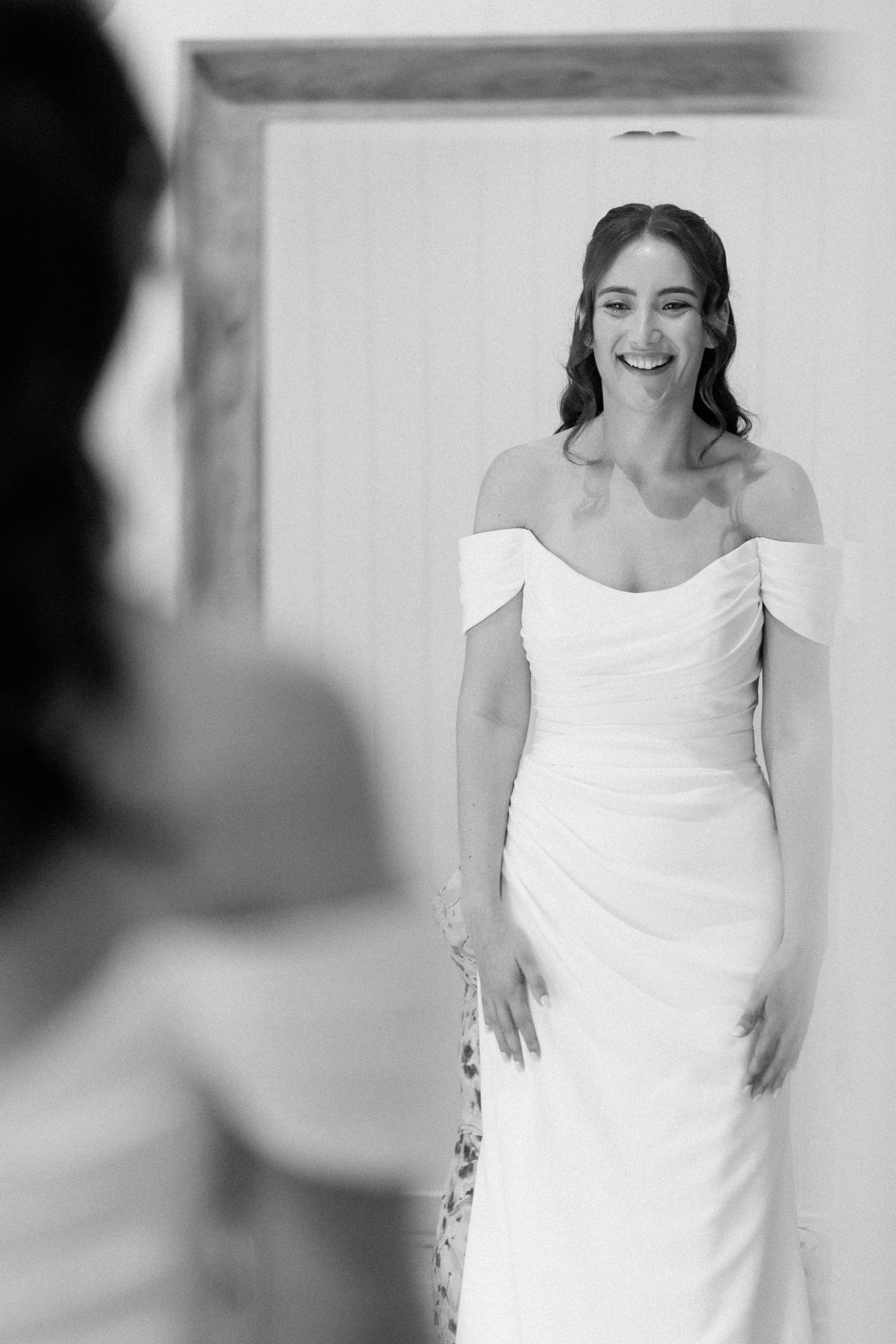 Bride in off-shoulder white gown smiling at her reflection in a mirror, black and white wedding photo.