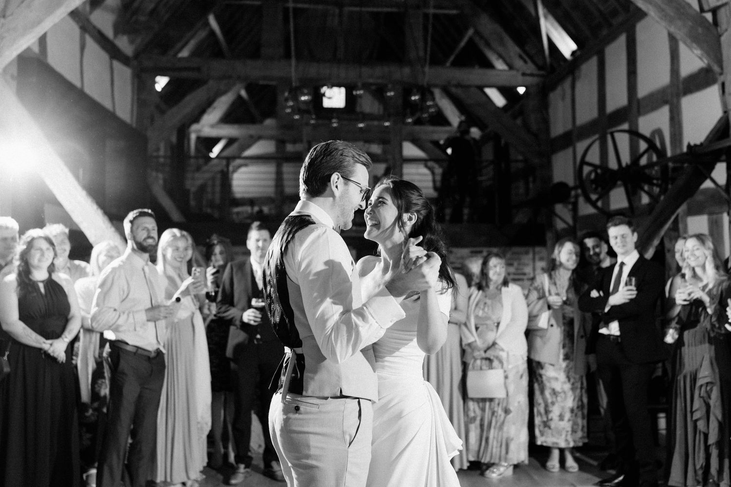 Black and white first dance photo of bride and groom in rustic barn surrounded by watching wedding guests.