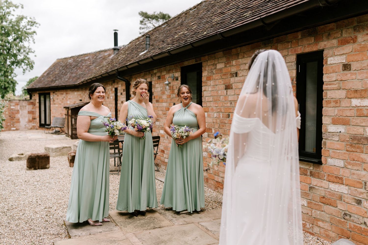 Bride in white veil faces three bridesmaids in sage green dresses holding bouquets outside a rustic brick venue.