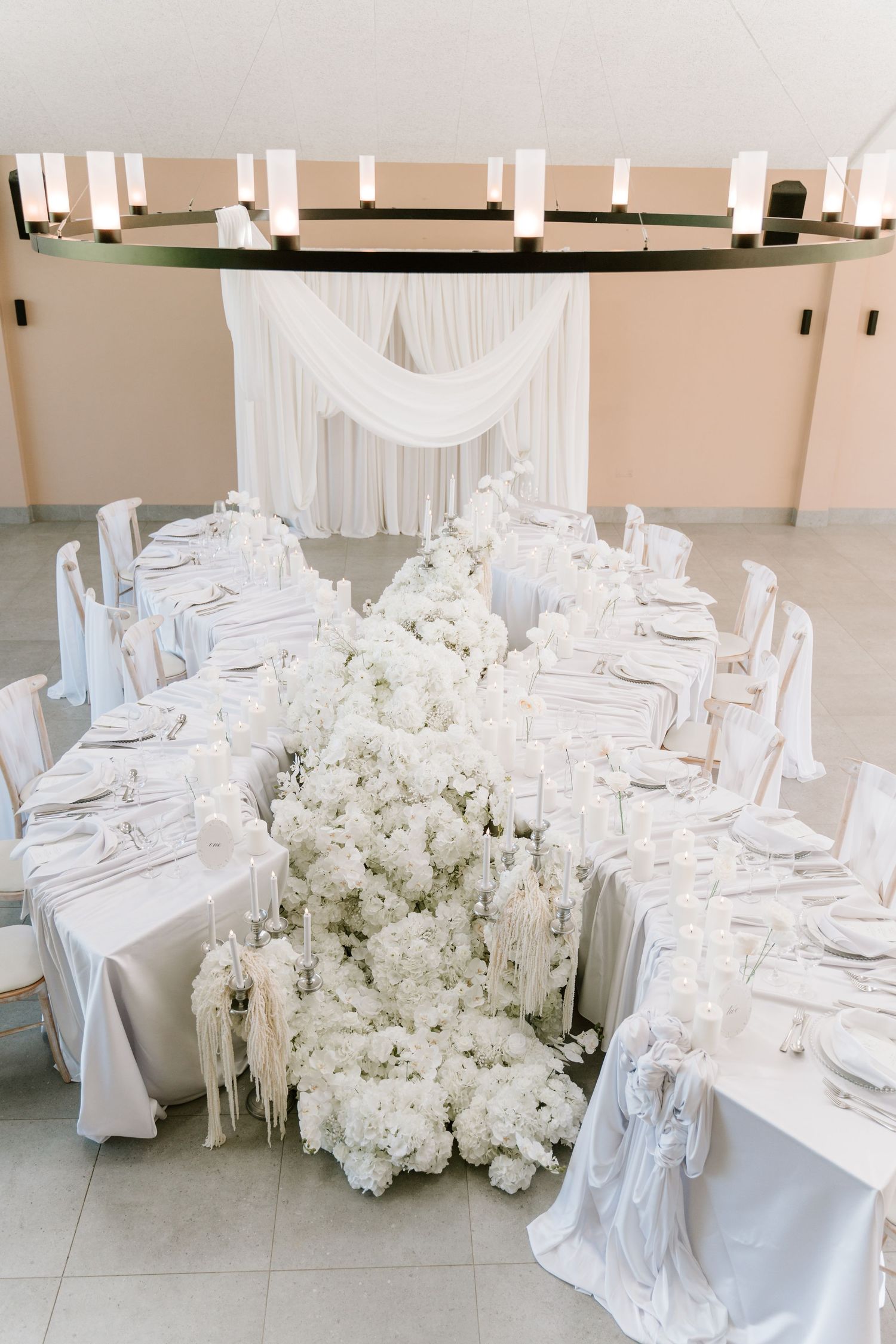 Elegant all-white wedding reception table adorned with lush floral arrangements, candles, and draped fabric backdrop.