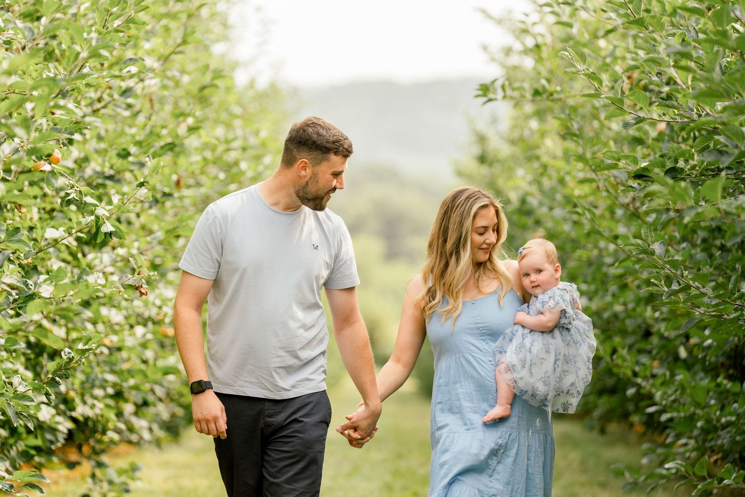A couple walks hand-in-hand through an apple orchard, the woman in a blue dress holding their baby.