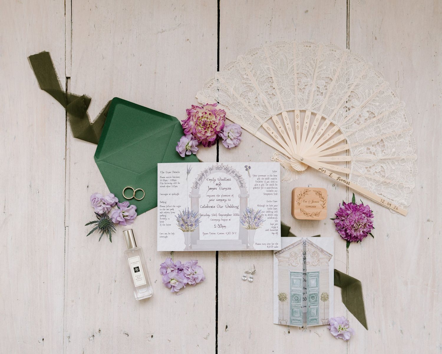 Elegant wedding flat lay with floral invitations, green envelope, lace fan, rings, perfume, and purple flowers on white wood.