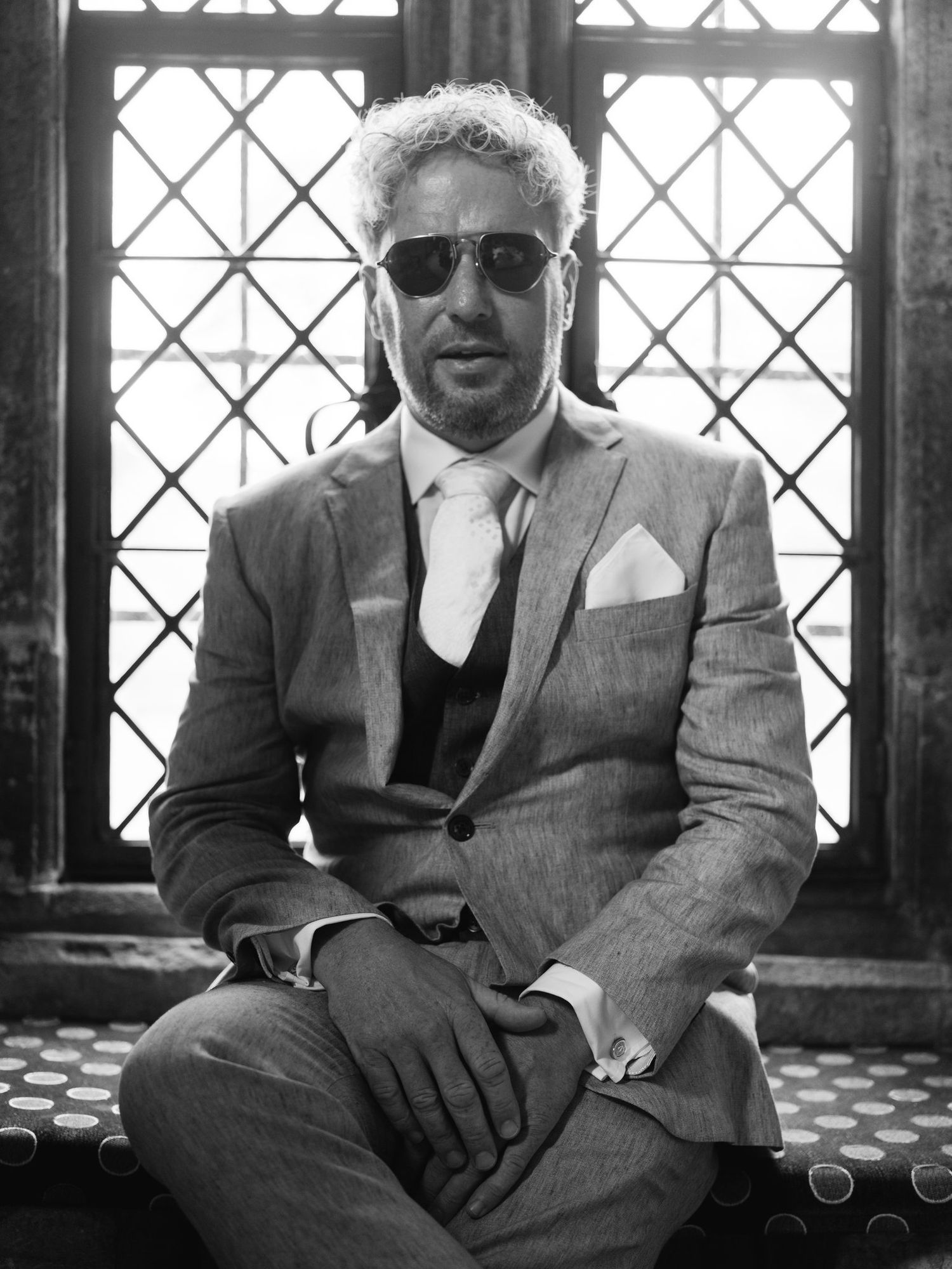 Groom in a three-piece suit and sunglasses by a leaded window at Brinsop Court