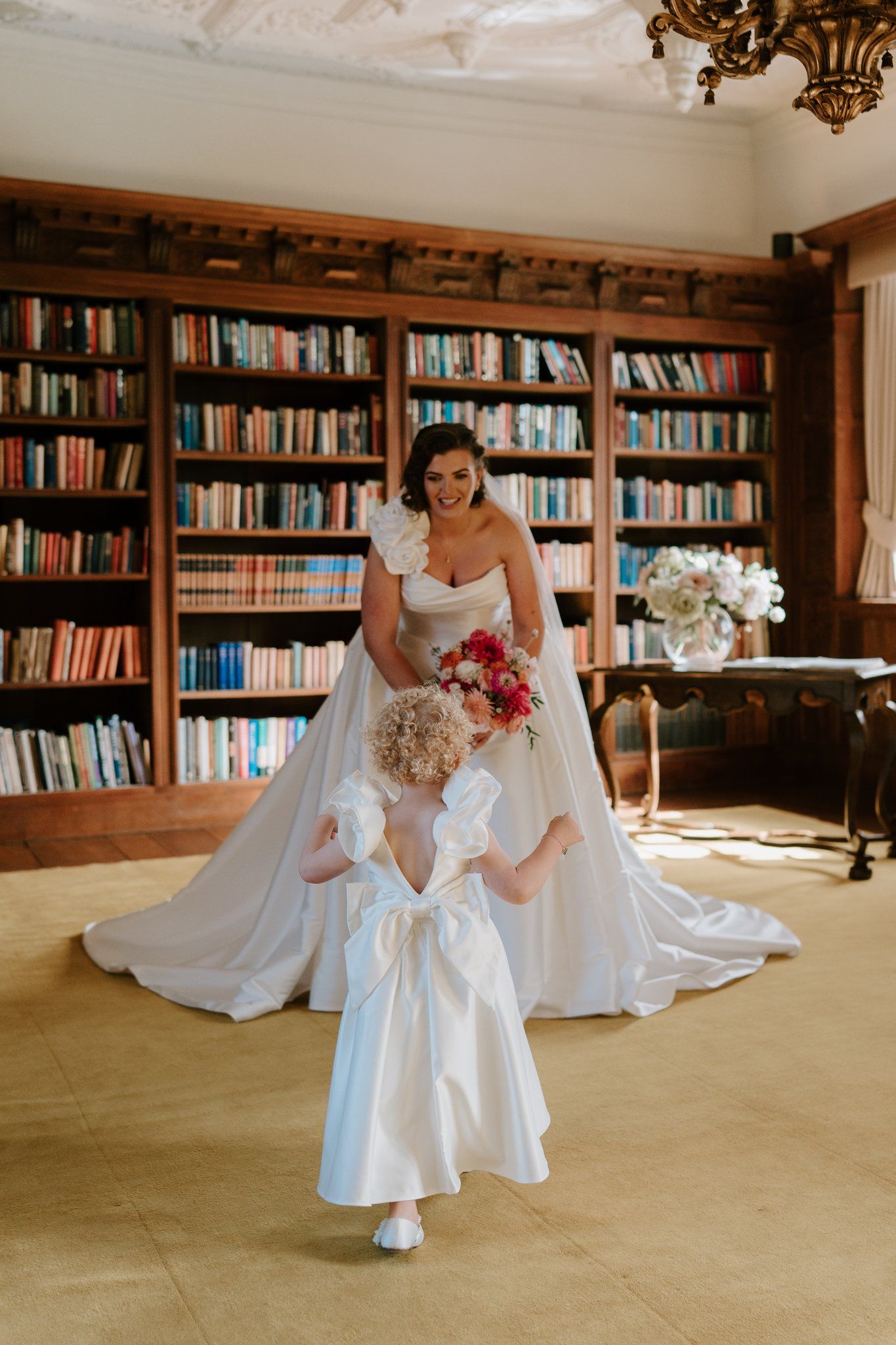 Bride in a strapless ball gown holds bouquet with flower girl in white dress in an elegant library setting.