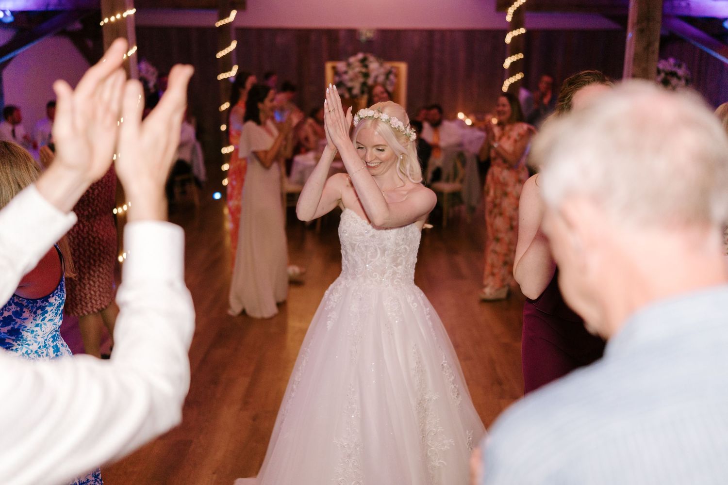 Bride in white gown dancing joyfully with guests at a wedding reception on a wooden dance floor.