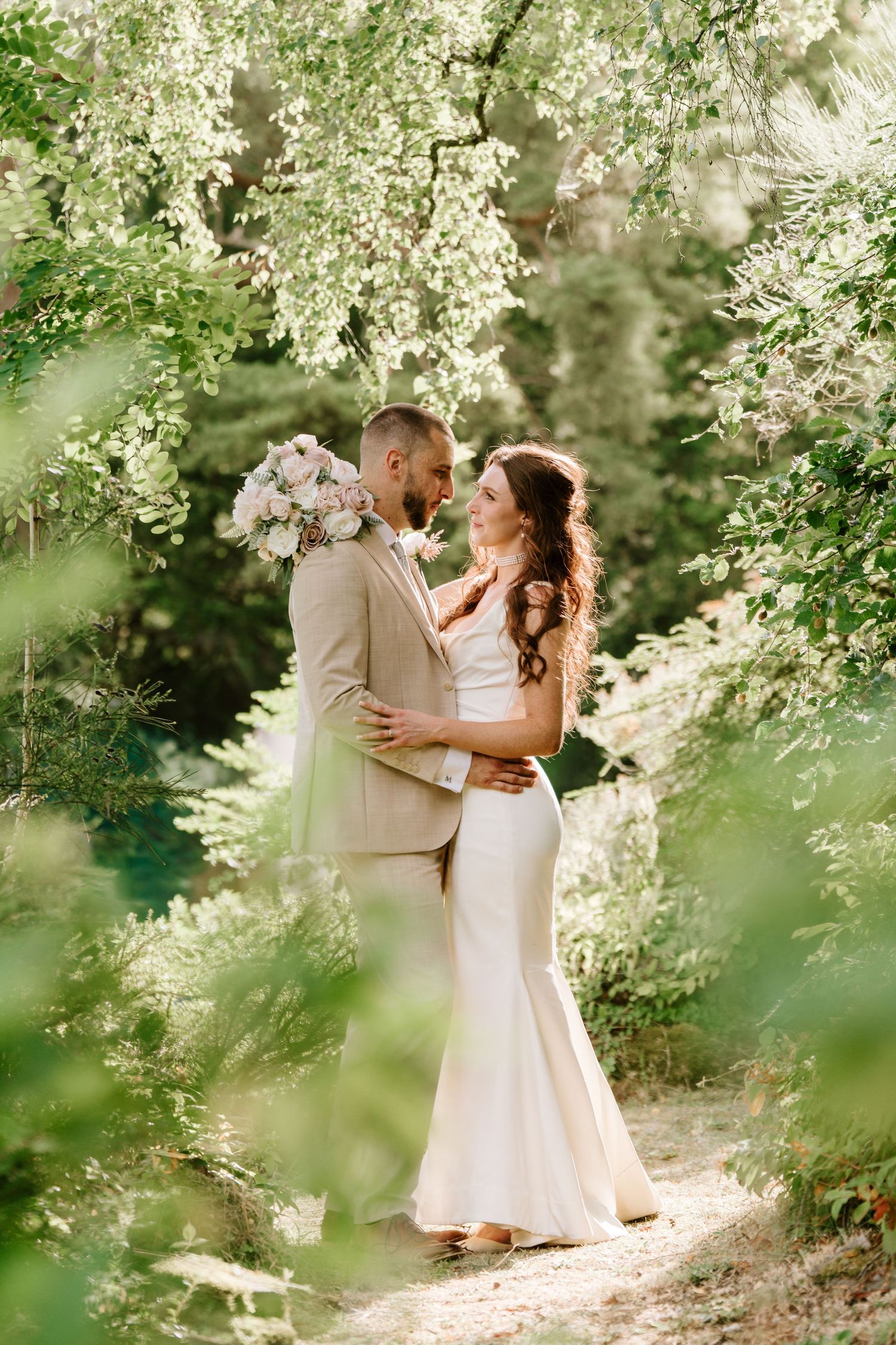 Wedding couple embracing under blooming trees in a sunlit garden, bride holding a pink bouquet.