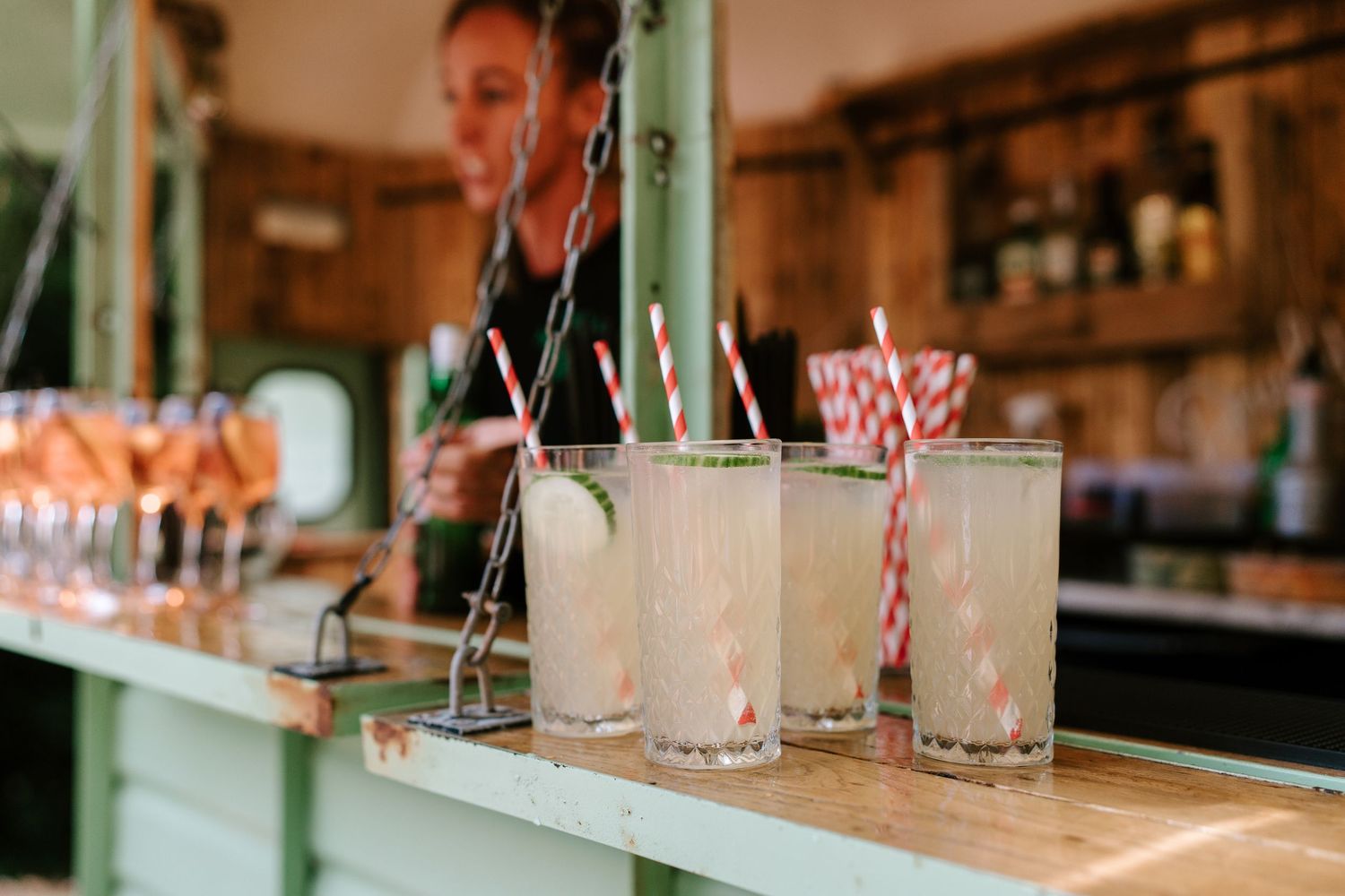 Cocktails with red striped straws on a bar counter with a bartender in the background.