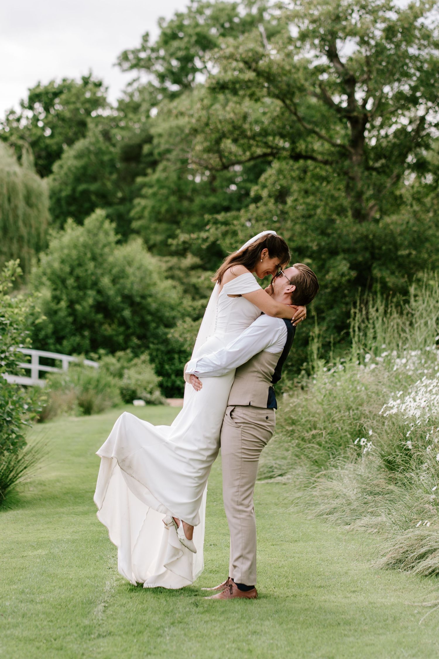 Groom lifting bride in white dress for a kiss in a lush green garden on their wedding day.