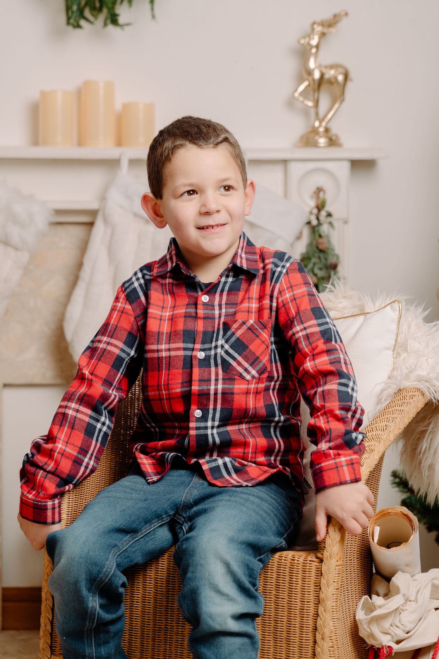 Young boy in red plaid shirt smiles while sitting in a wicker chair in a cozy Christmas decorated room.
