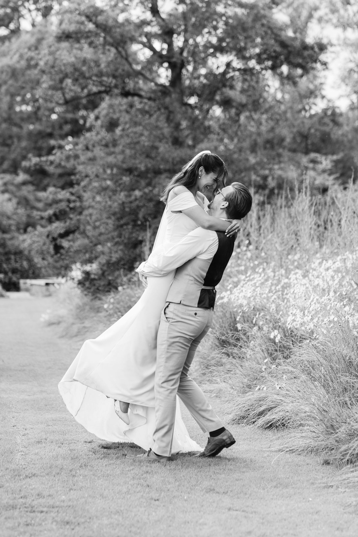 Groom lifts bride in a romantic black and white wedding photo surrounded by lush greenery and wildflowers.