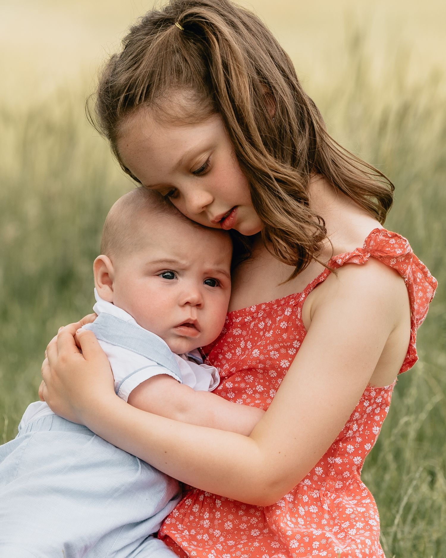 A young girl in a red floral dress tenderly hugs a baby in a sunlit green field.