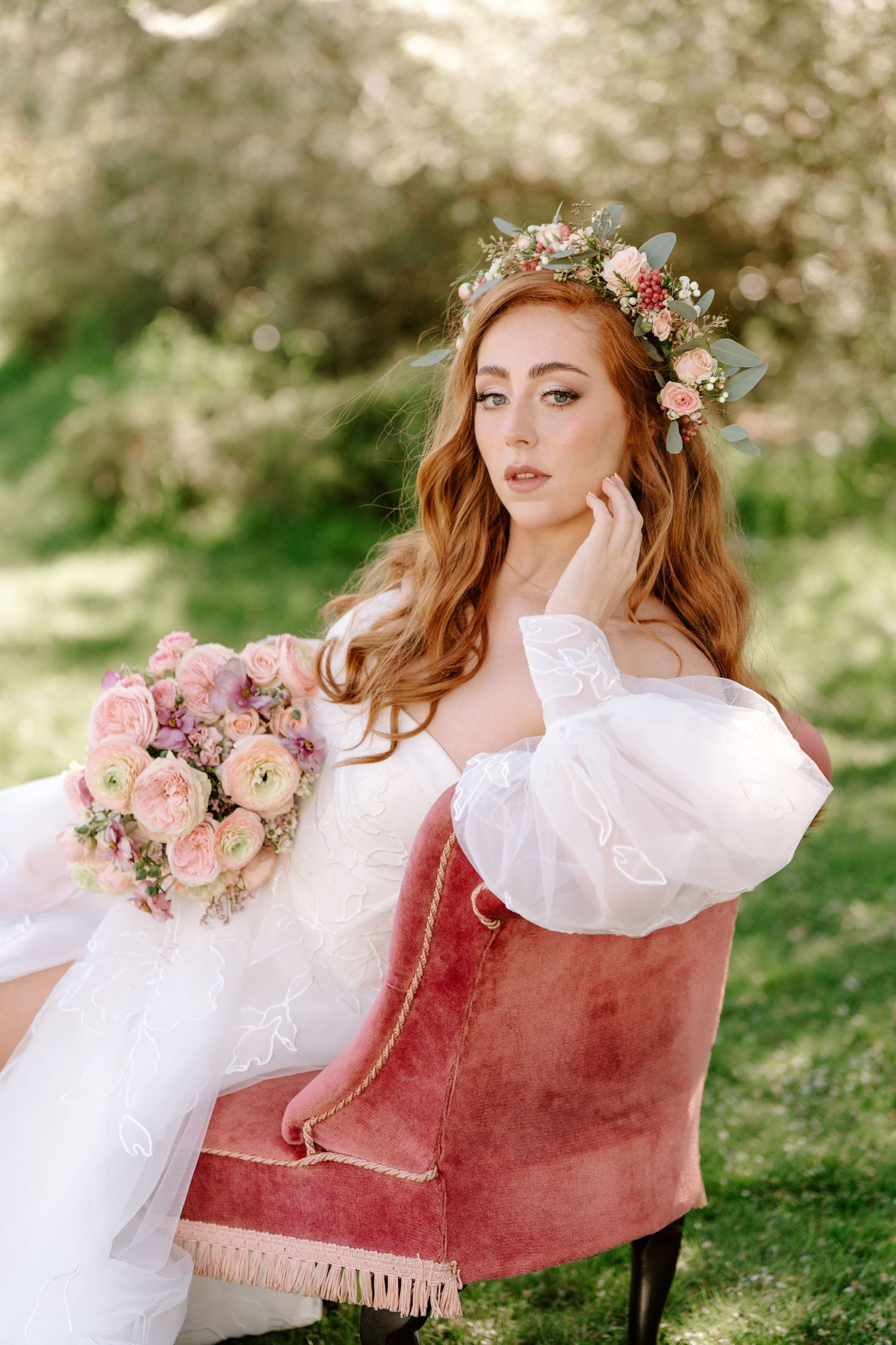 Red-haired bride in white gown with floral crown holds pink bouquet, seated on velvet chair outdoors.