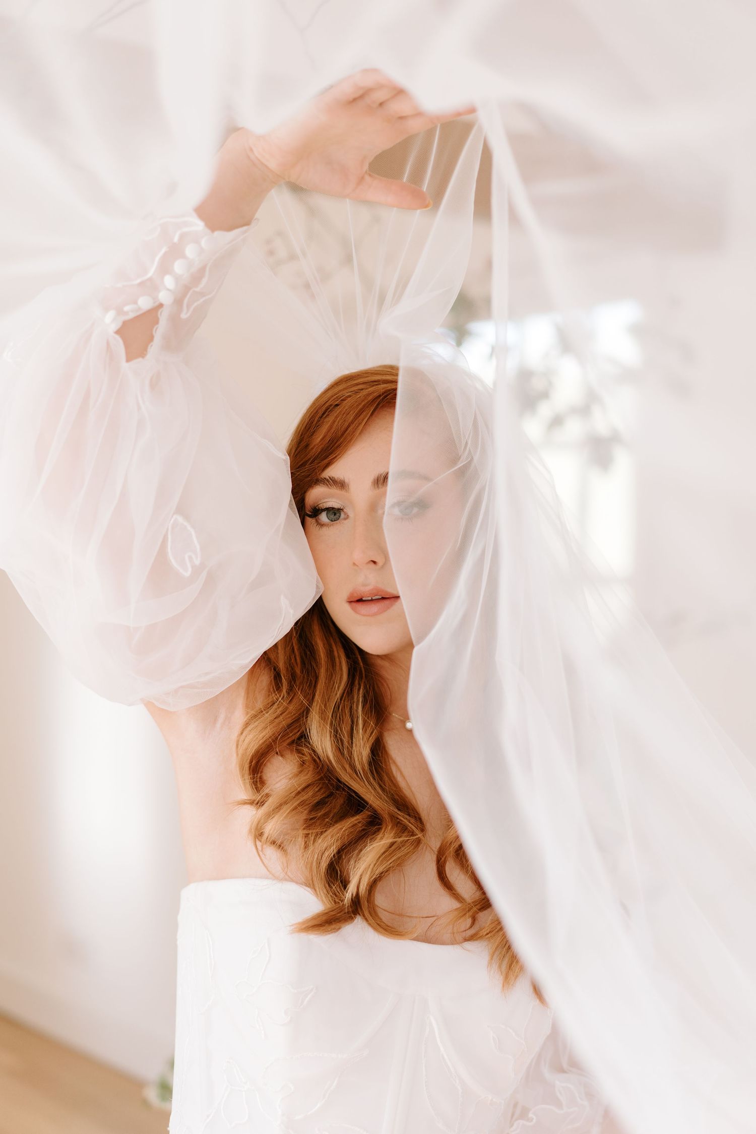 A red-haired bride lifts her sheer white veil, wearing an off-shoulder gown in a softly lit bridal suite.
