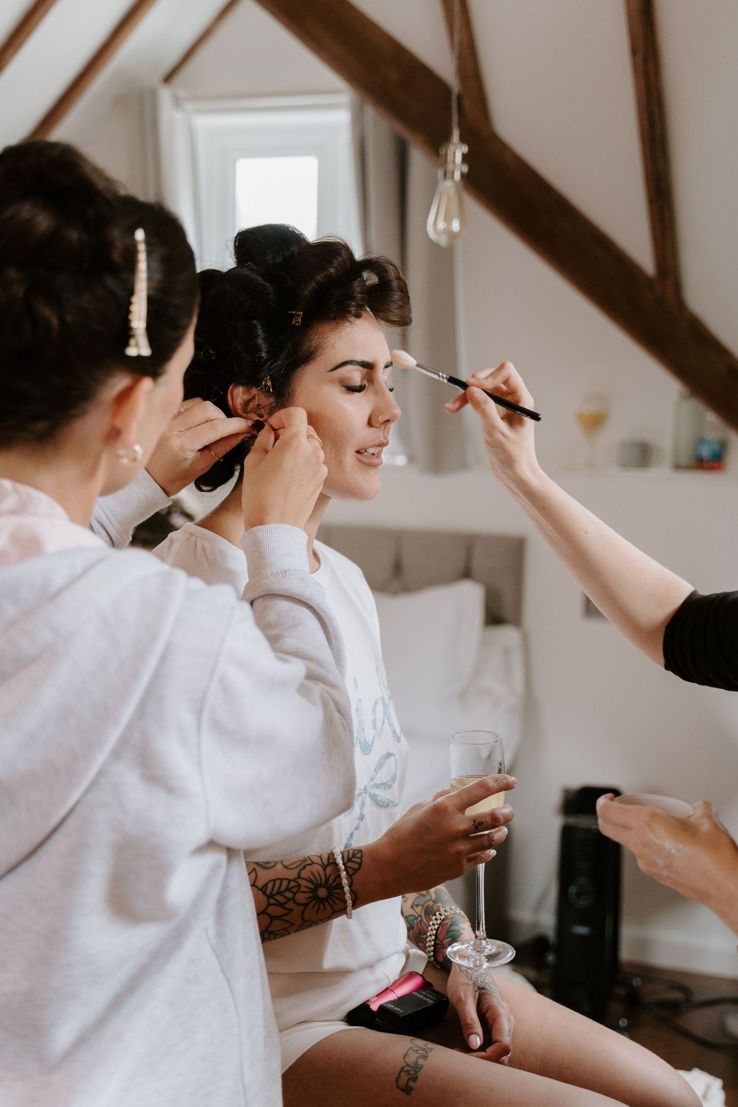 Bride getting makeup and earrings put on by bridesmaids in a cozy attic room before her wedding.
