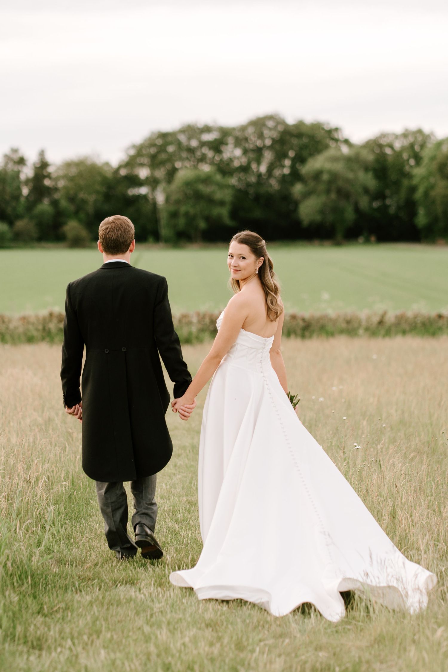 Bride in white gown looks back smiling while holding groom's hand walking through green field.