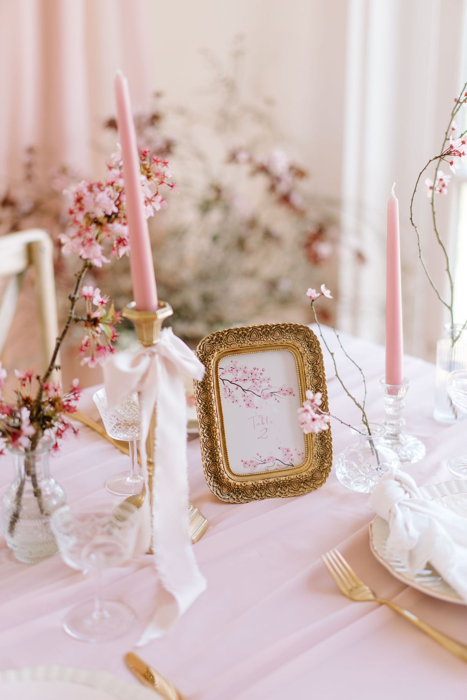 Elegant pink wedding table setting with gold candlesticks, cherry blossom florals, gold flatware, and a framed table number card.