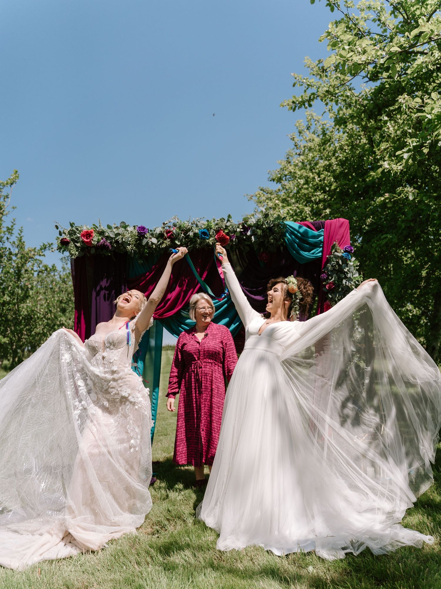 Two brides in white gowns celebrate outdoors with a floral chuppah and colorful fabric backdrop on a sunny wedding day.