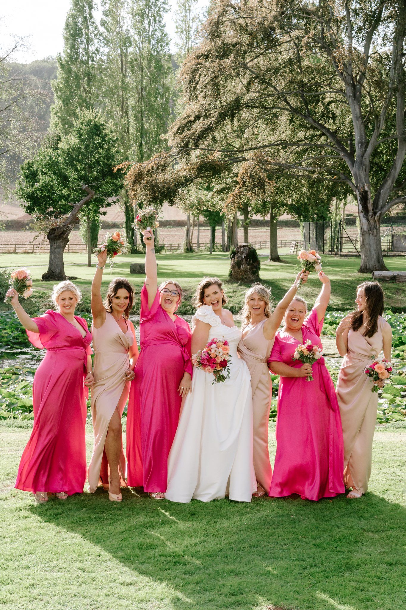 Bride in white gown poses with six bridesmaids in pink and blush dresses, raising bouquets outdoors.