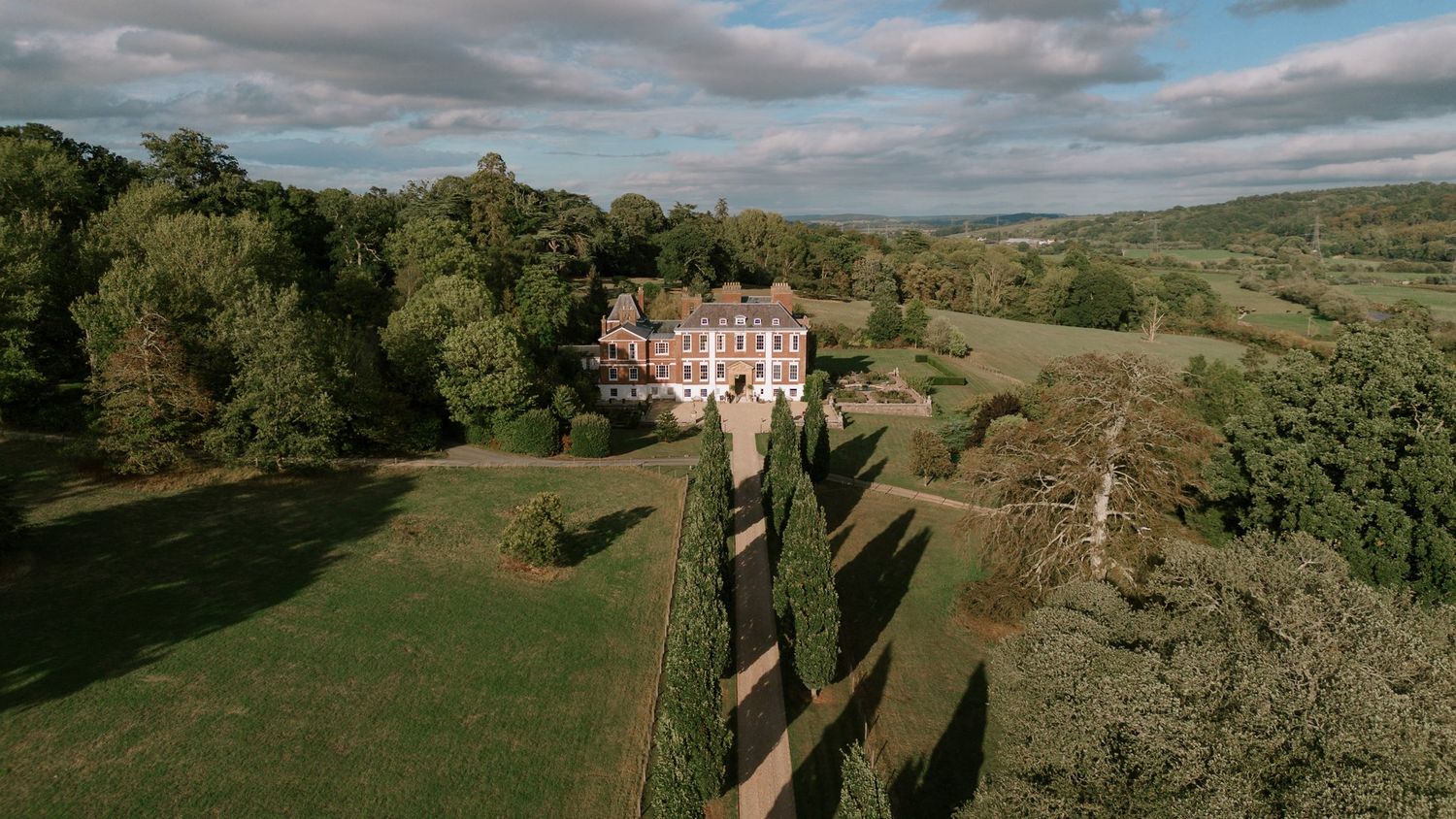 Aerial view of a grand English country manor surrounded by manicured hedges and lush green countryside under a cloudy sky.