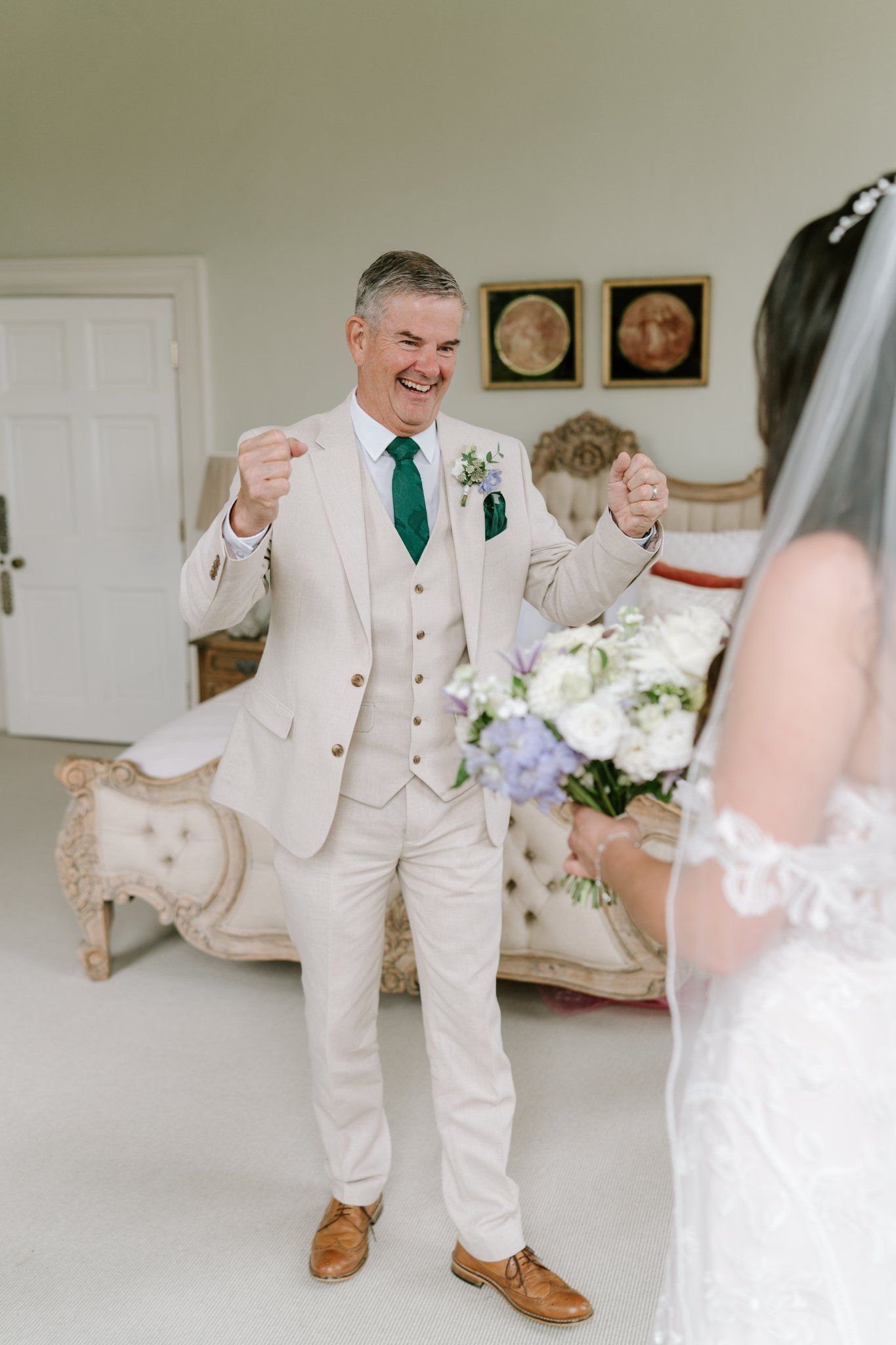 Groom in cream suit with green tie reacts with joy seeing bride in white lace wedding dress holding bouquet indoors.