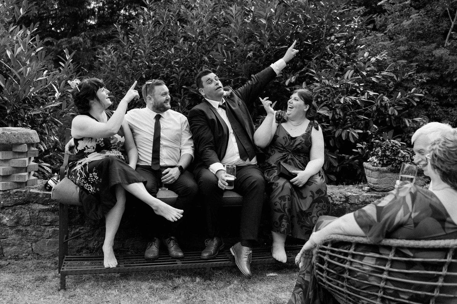 Wedding guests laughing and relaxing together on stone bench in garden in black and white.