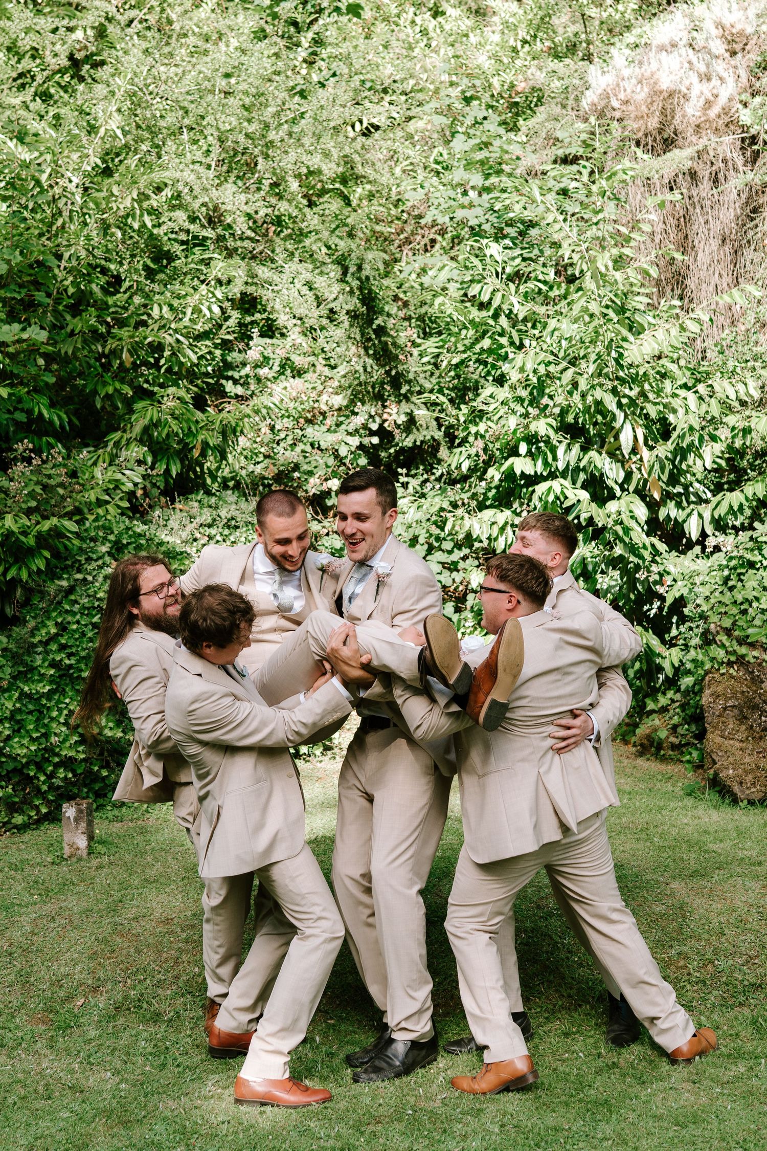 Groomsmen in tan suits playfully lifting the groom outdoors in a lush green garden during a wedding celebration.