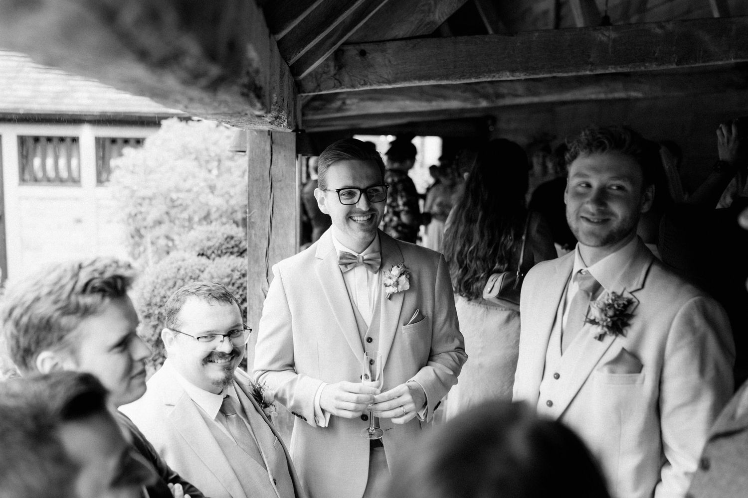 Groom in light suit and bow tie laughing with groomsmen at outdoor wedding ceremony in black and white.