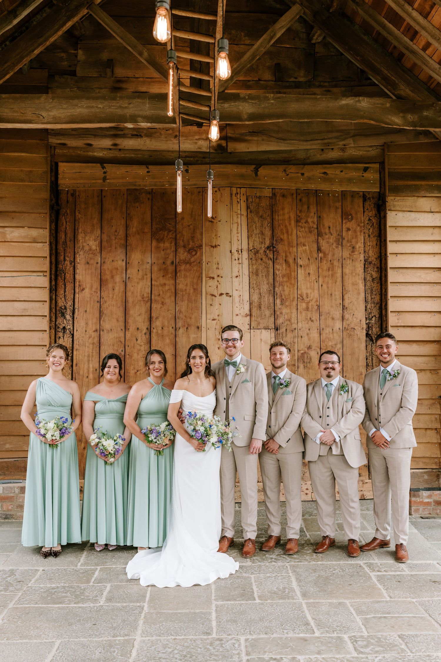 Wedding party posing before rustic wooden barn doors, bridesmaids in sage green and groomsmen in tan.