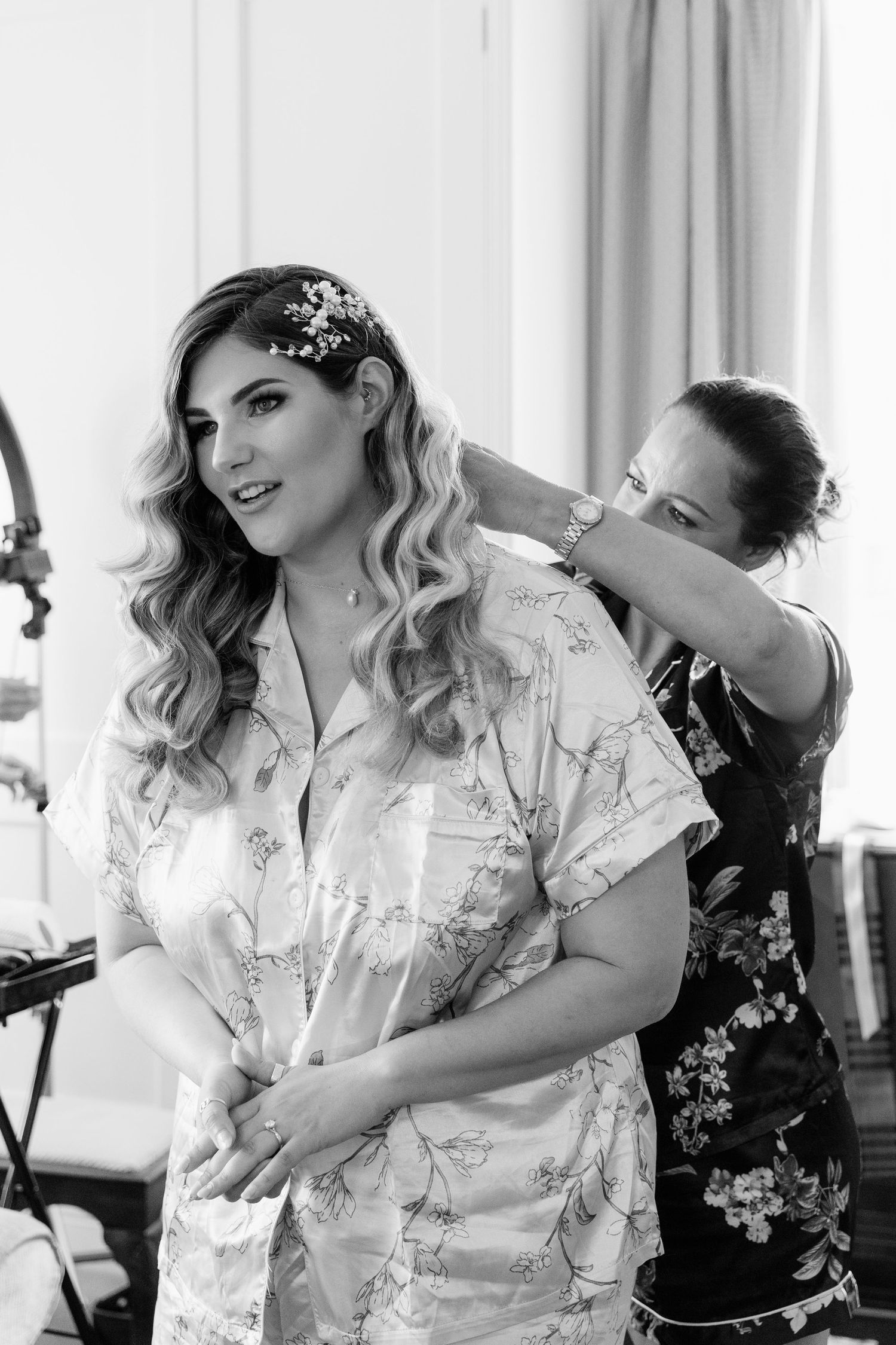 Bride in floral robe gets hair accessory placed by bridesmaid during wedding preparations, black and white photo.