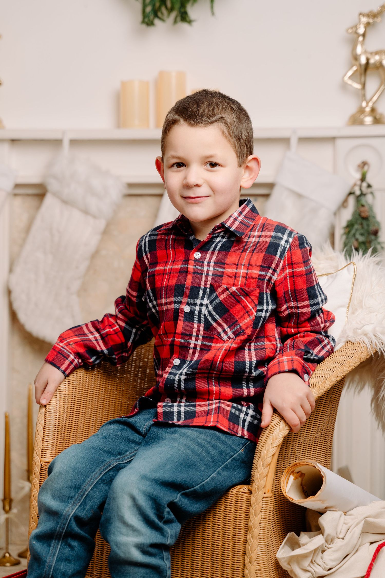 Young boy in red plaid shirt and jeans sitting in wicker chair by Christmas fireplace with stockings.