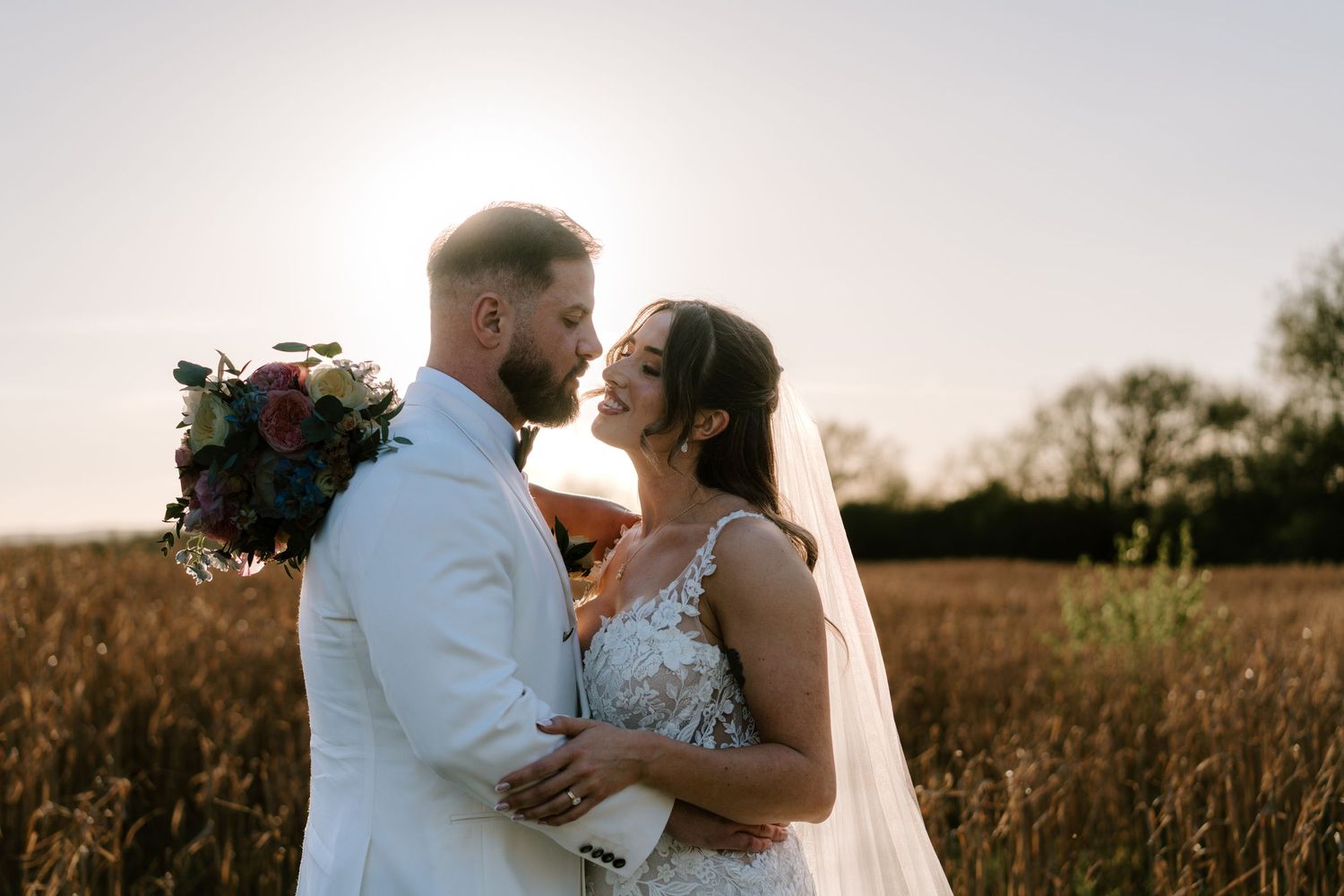 Bride and groom share a romantic moment in a golden field at sunset, bride holding a burgundy bouquet.
