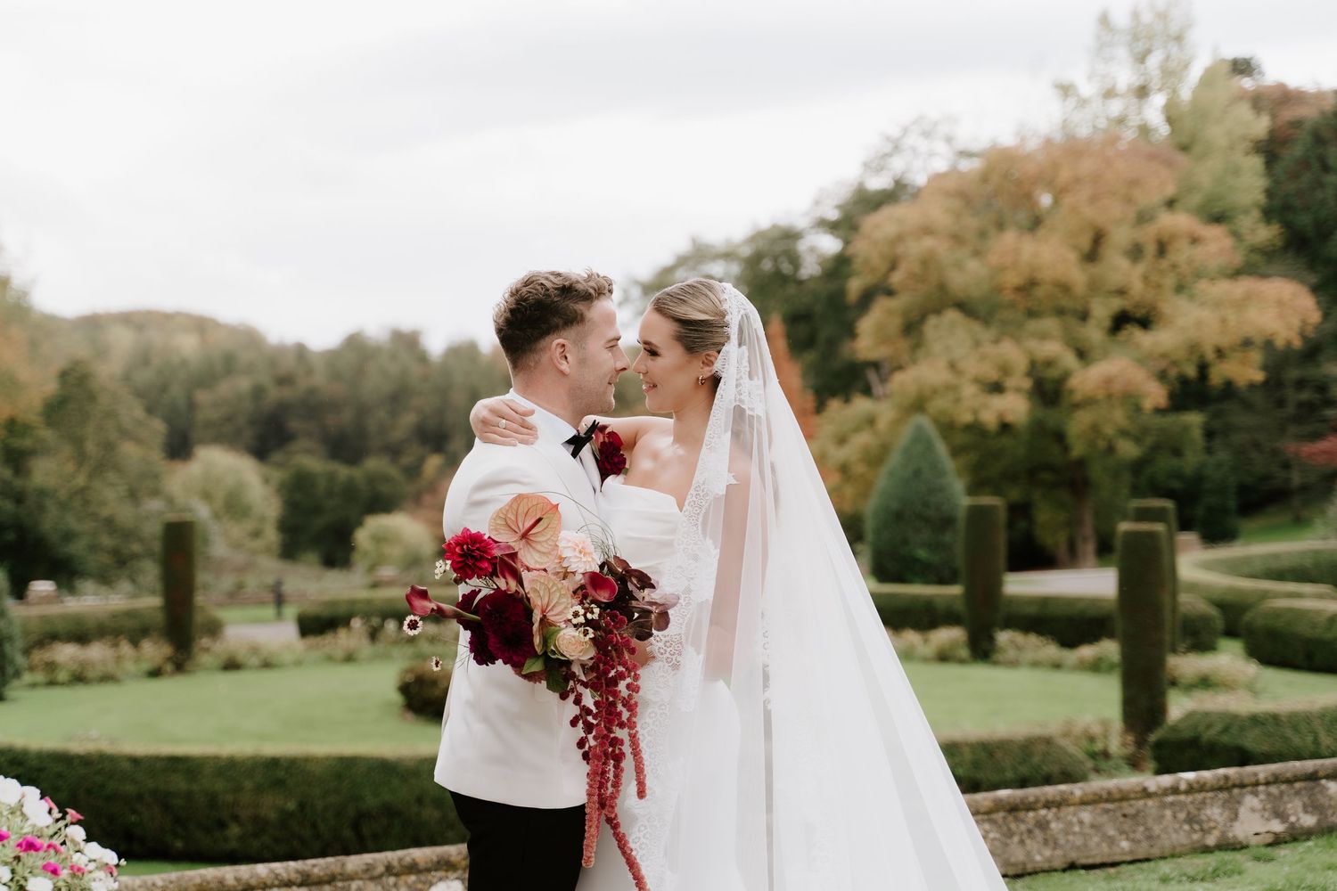 Bride and groom sharing a romantic kiss in a lush garden, bride holding a red bouquet and wearing a flowing veil.